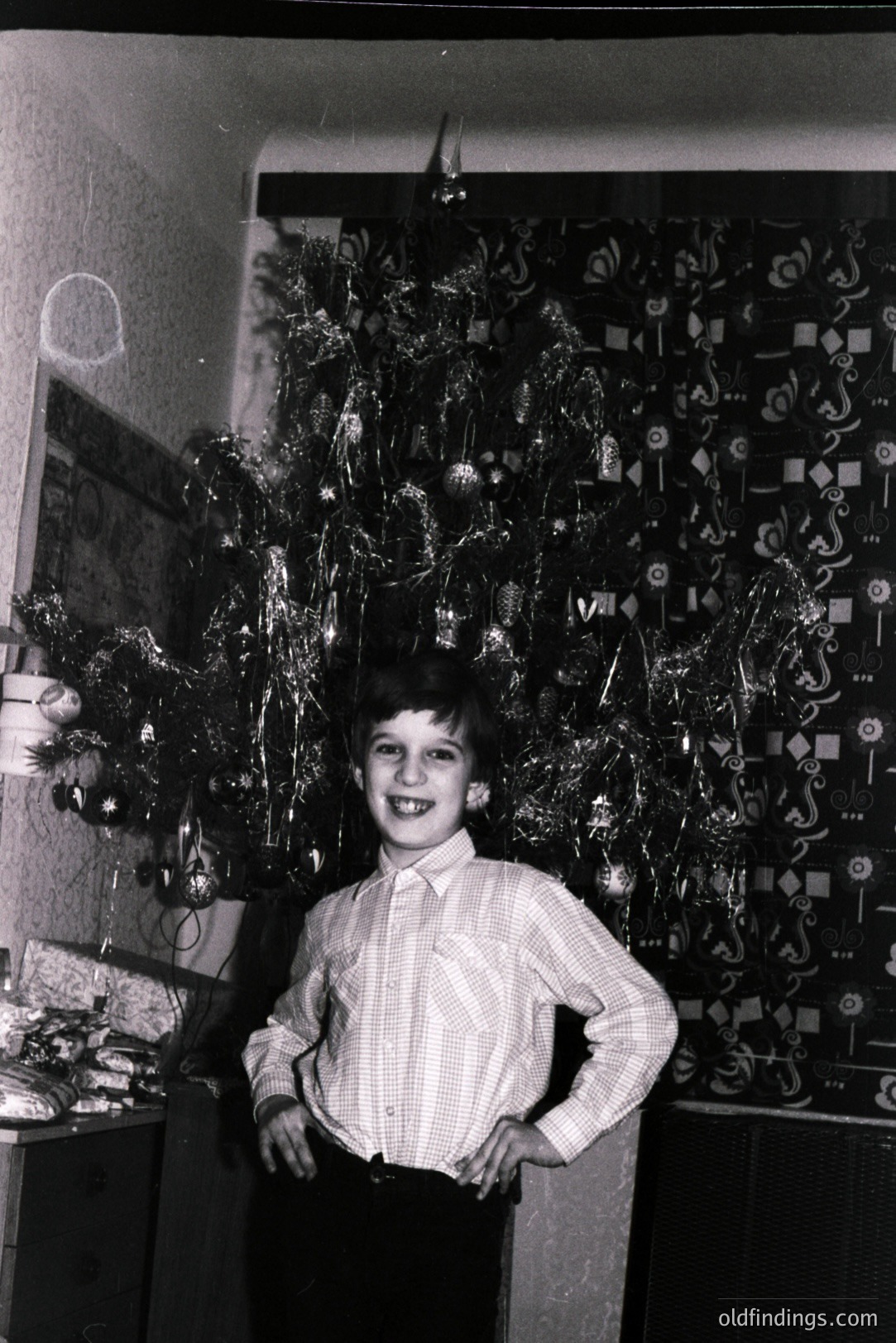 A young boy stands proudly with hands on hips in front of a tinsel-covered Christmas tree. The room’s patterned wallpaper and vintage furniture suggest a mid-century interior. Likely taken in the 1960s or 1970s, it offers a nostalgic glimpse into a family holiday.