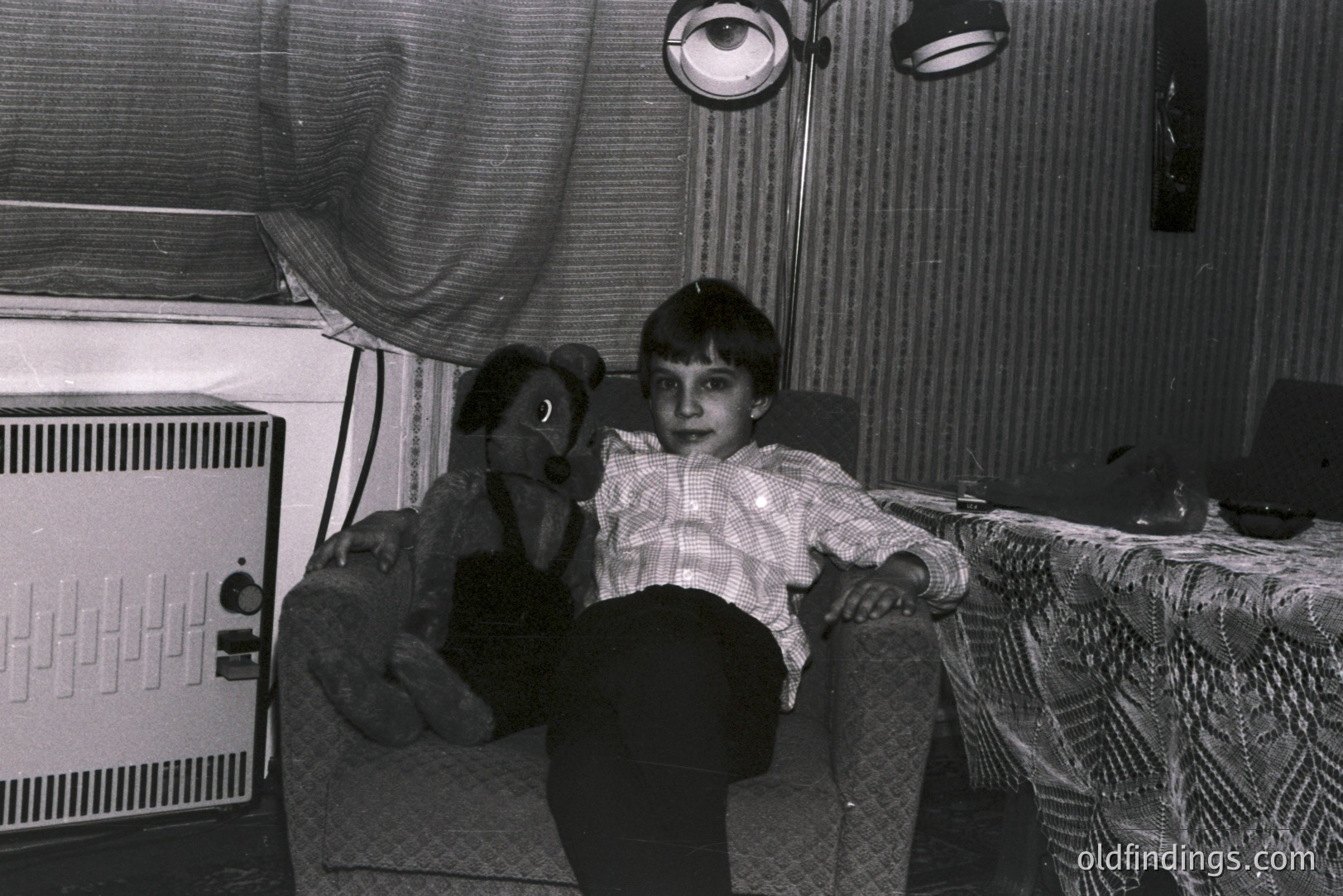 A young boy sits on a patterned armchair, alongside a worn teddy bear. He wears a button-down shirt & dark pants. A textured curtain & vintage lamp suggest a domestic interior, possibly 1960s-70s. The image shows a candid, intimate moment.