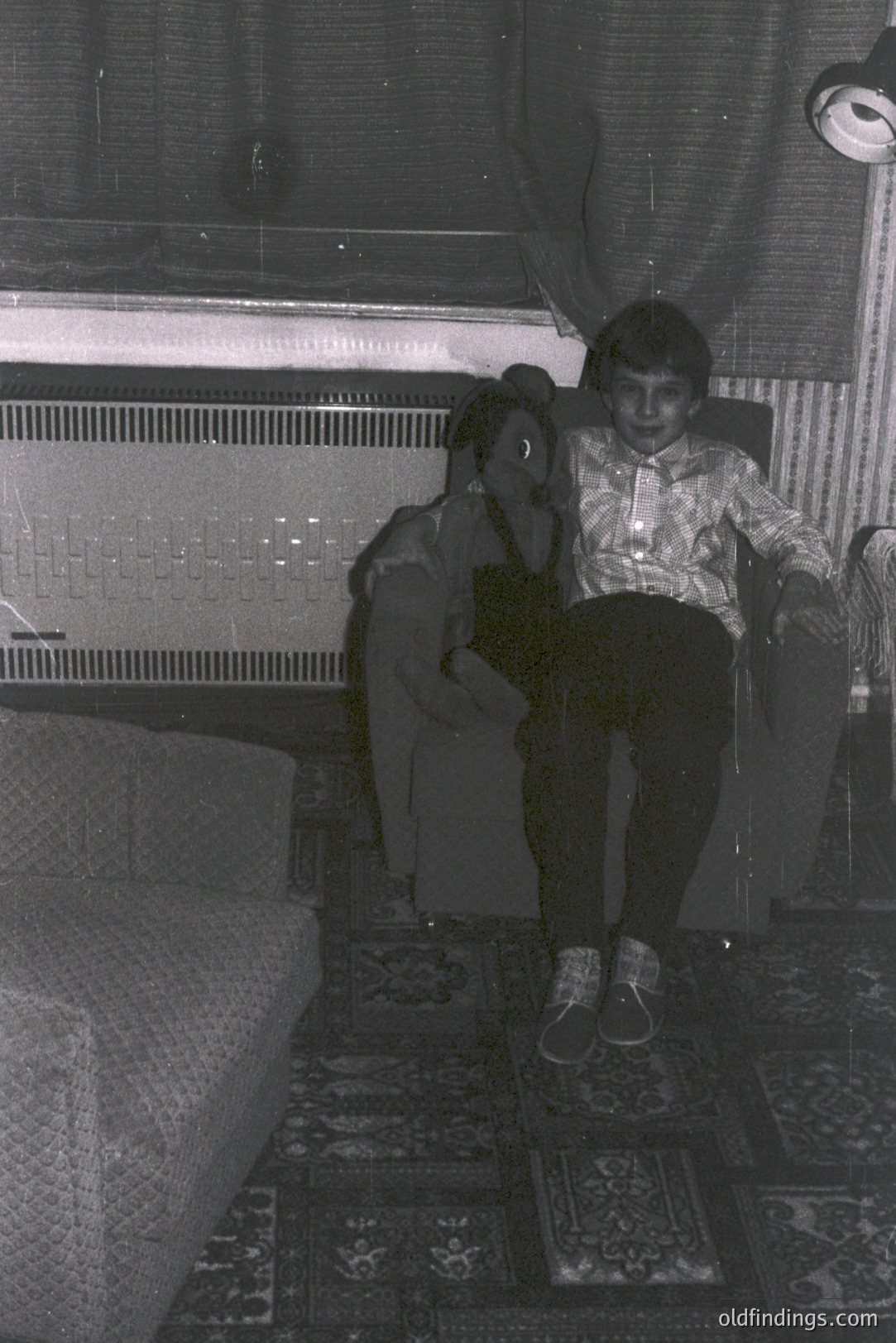 A young boy and girl sit together in a richly patterned room. The boy, wearing a patterned shirt and dark trousers, is seated on a cushioned chair. The girl is partially obscured, wearing a sweater. Vintage style decor and a detailed rug suggest a mid-century interior.