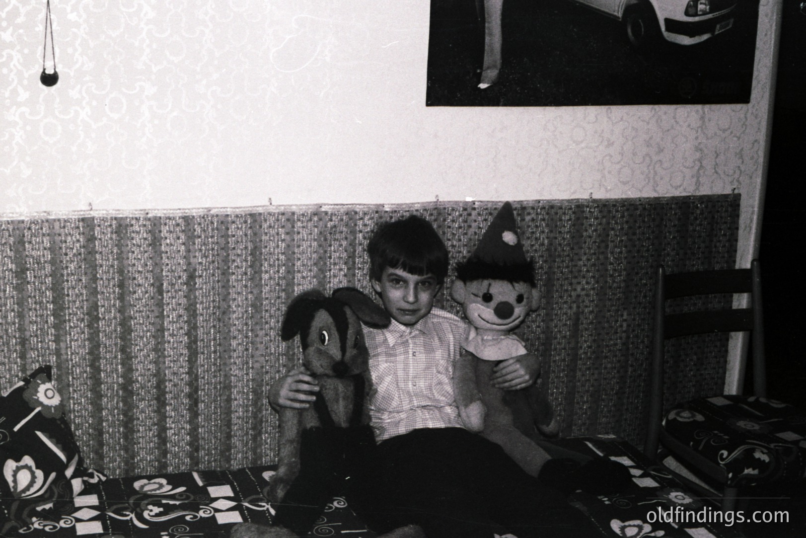 A young boy sits on a patterned sofa, holding a stuffed dog and clown. Visible décor suggests a modest, likely European interior, circa 1960s-70s. The image's simplicity lends itself to nostalgic portraiture or design references. Photo evokes childhood and domestic life.