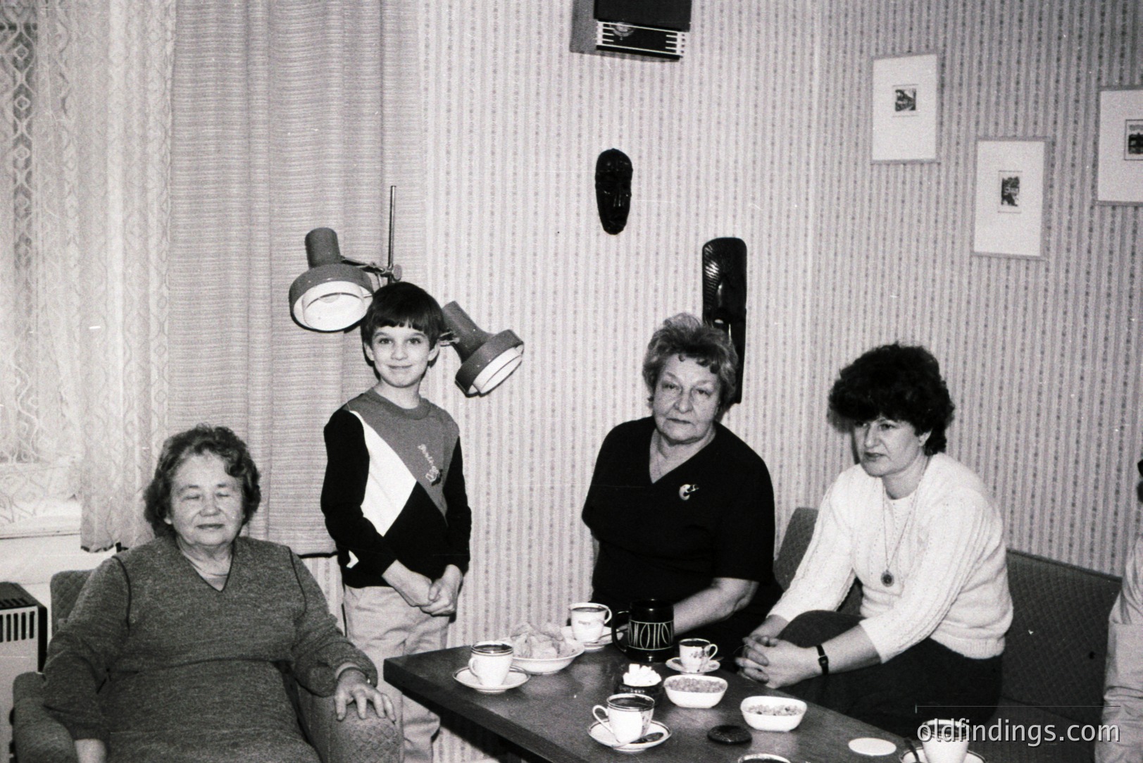 Four individuals are captured in a snapshot, likely a family portrait from the 1970s. A young boy stands beside a woman seated on a plush chair, while two more women are positioned at a table set with teacups. The interior features distinctive 70s decor – textured wallpaper and a large floor lamp.