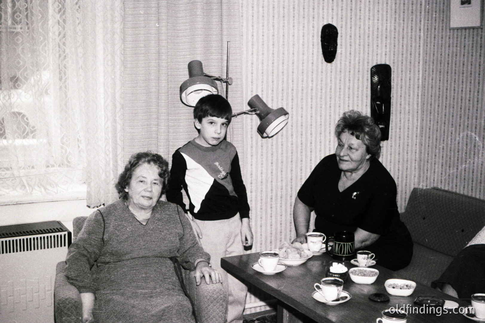 A candid black and white portrait captures a young boy between two women, seemingly at a table set for tea. Architectural details suggest a mid-century interior with vertical patterned wallpaper. The room's lighting is provided by a distinctive double-armed lamp.