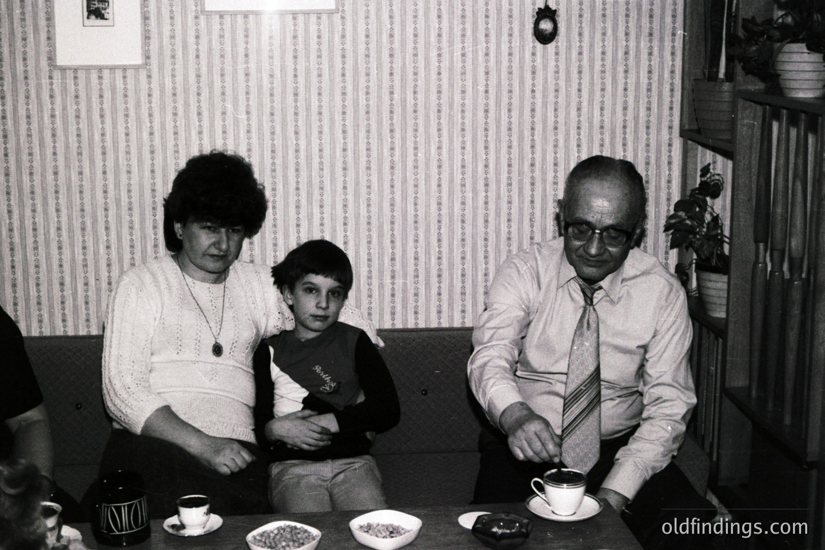 A family portrait, likely 1970s. A grandmother, boy, and grandfather are seated at a table with floral patterned wallpaper in the background. The man wears glasses & a striped tie; the boy has short hair. A coffee service & sweets are on the table.