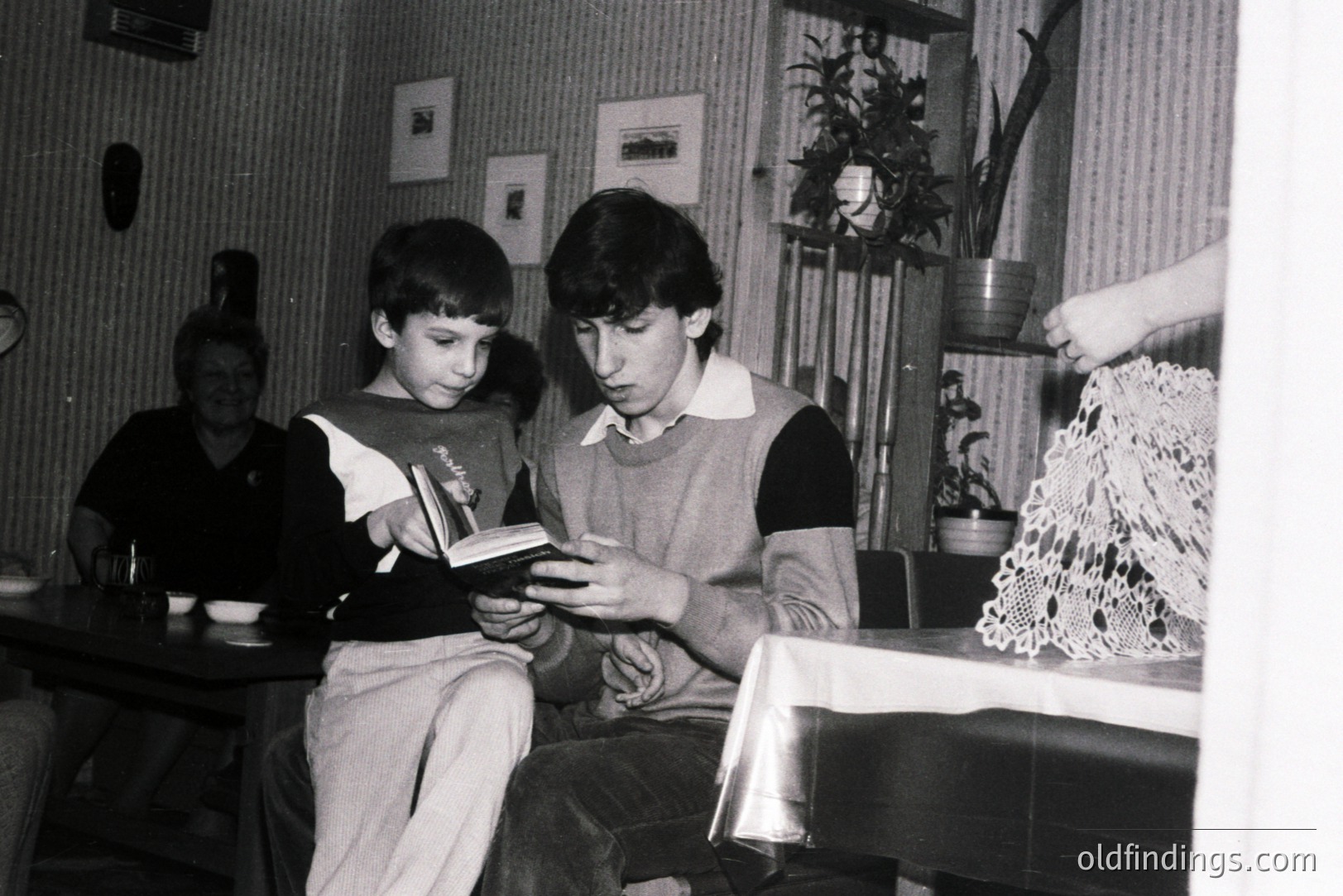 Two boys intently examine a book seated at a table draped with lace. A third person is visible in the background. Likely a domestic interior, possibly a restaurant or cafe. The style suggests a 1970s or 1980s aesthetic.