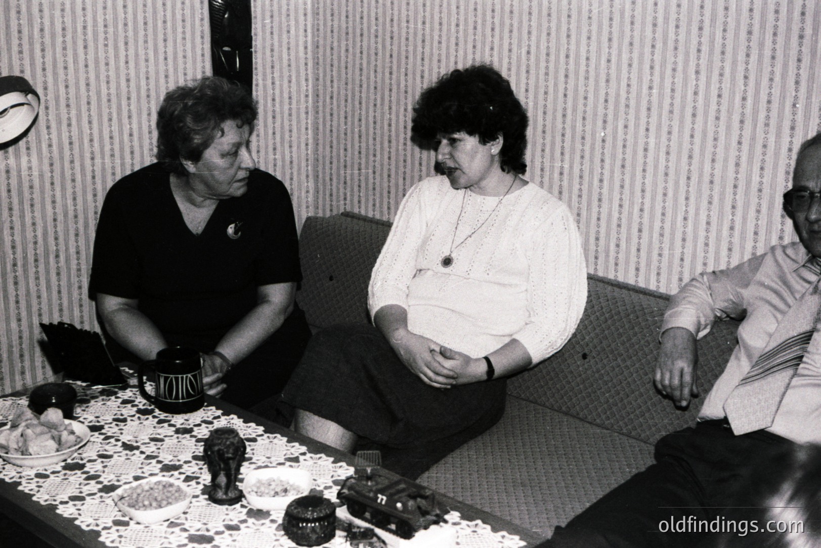 A candid, black and white photograph depicts three individuals engaged in conversation within a residential setting. Two women sit across a table, while a man sits on a nearby sofa. The table displays decorative items and a branded container. Interior suggests a 1970s or 1980s home.