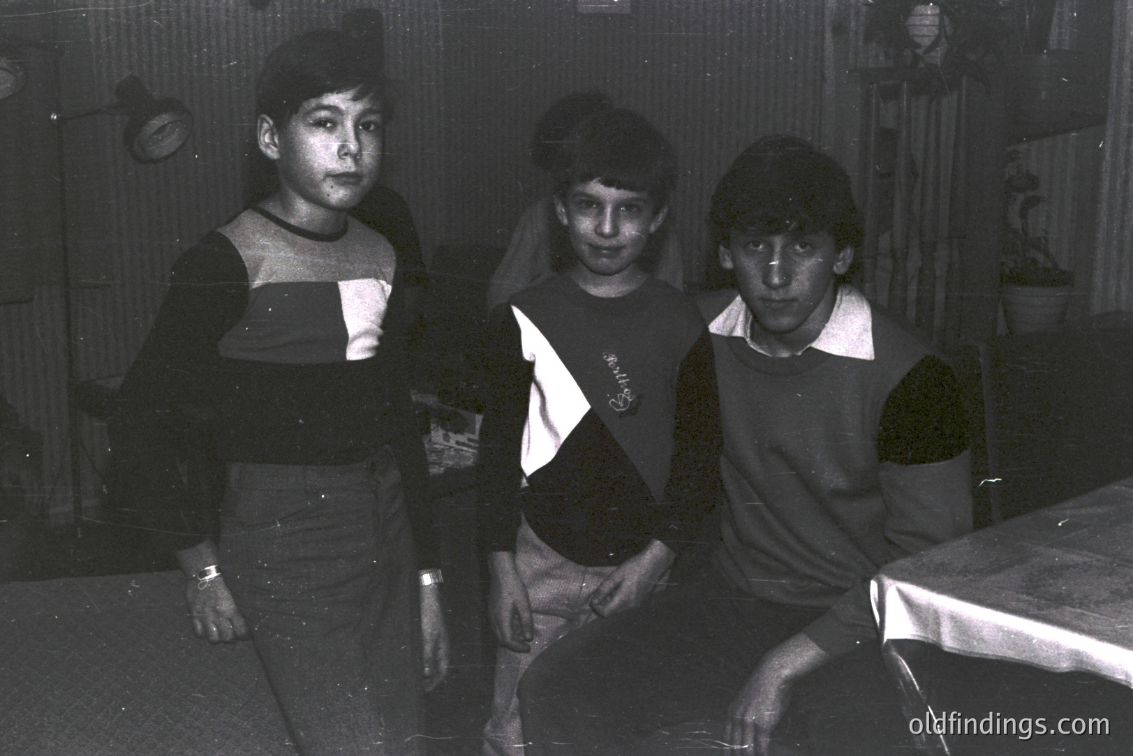 Three young boys in casual, dark-colored sweaters and pants pose in a home interior. A table covered with cloth is visible. Likely a family snapshot; the décor suggests a modest, 1970s-era domestic setting. Photo’s quality indicates amateur photography.