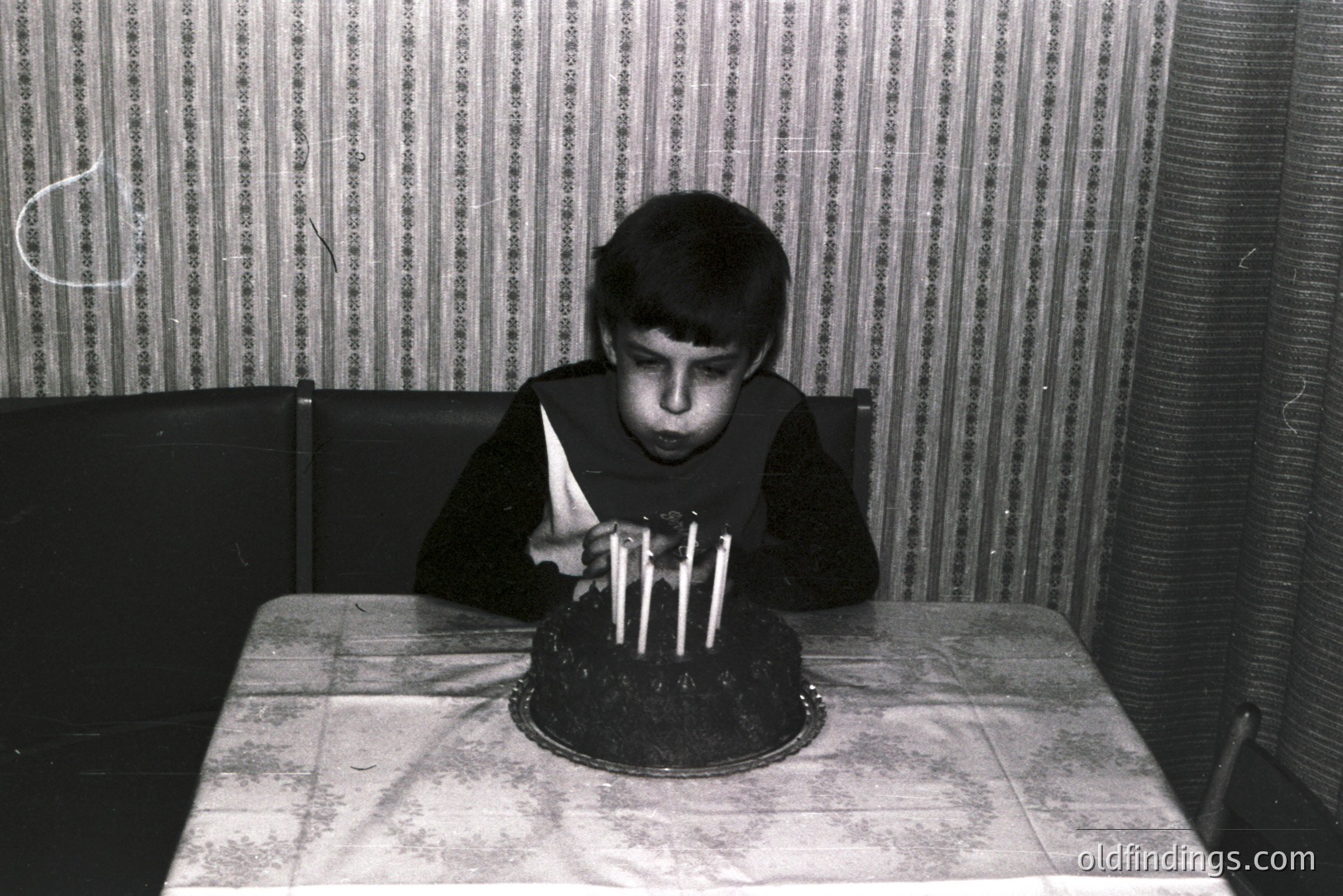 A young boy blows out four candles on a dark chocolate cake covered in white frosting. He holds a folded napkin and is seated at a table draped with a thin fabric. Background features textured wallpaper & dark furniture. Likely a candid, family portrait, circa 1960s-1970s. Simple composition, appealing for vintage design.