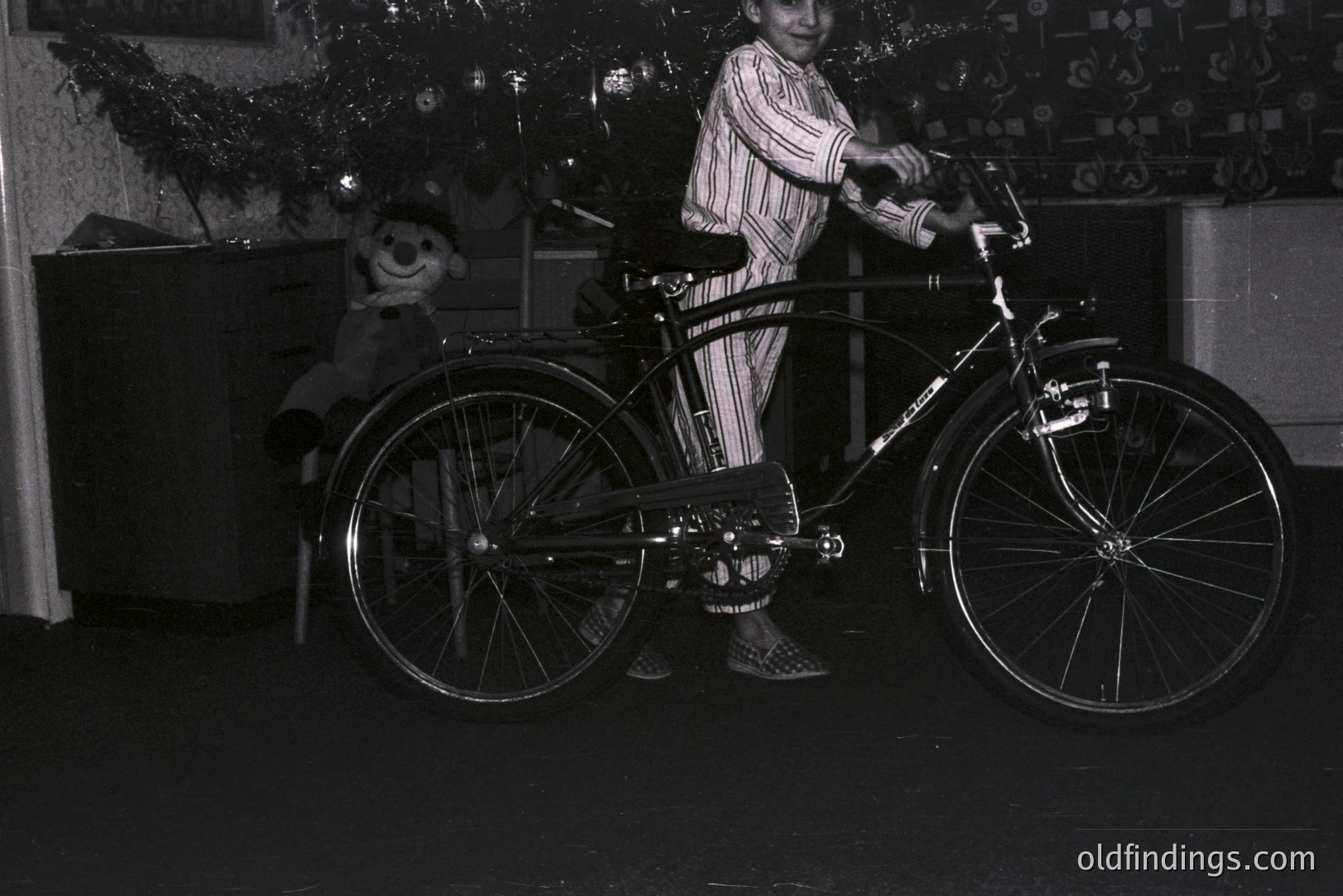 A young boy in striped pajamas stands proudly beside a vintage Schwinn bicycle, likely a gift. A teddy bear sits nearby. Decorations suggest a holiday scene, possibly Christmas. The bicycle’s design, with fenders and a front rack, is characteristic of mid-20th century American bicycles.