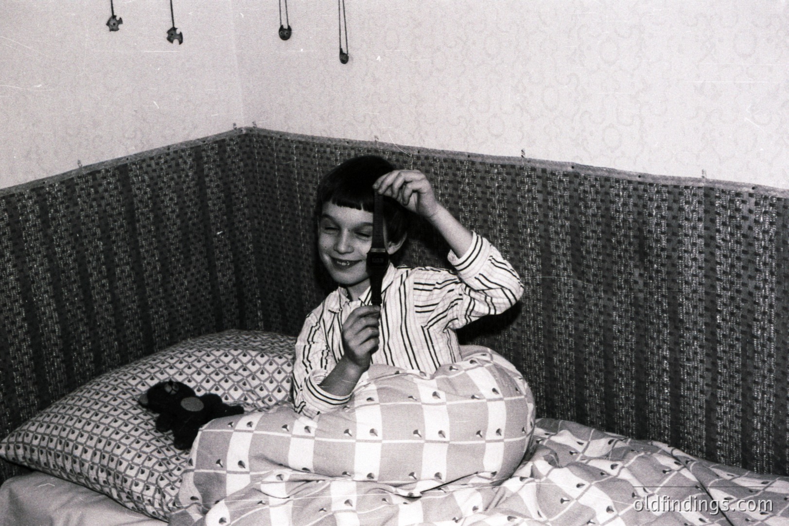 A young girl, likely a child, lies in a bed with a woven headboard. She wears striped pajamas and playfully holds a brush to her hair while looking at the camera. The room has a textured wallpaper backdrop and a simple, mid-century aesthetic. Possibly a snapshot from the 1960s or 70s.