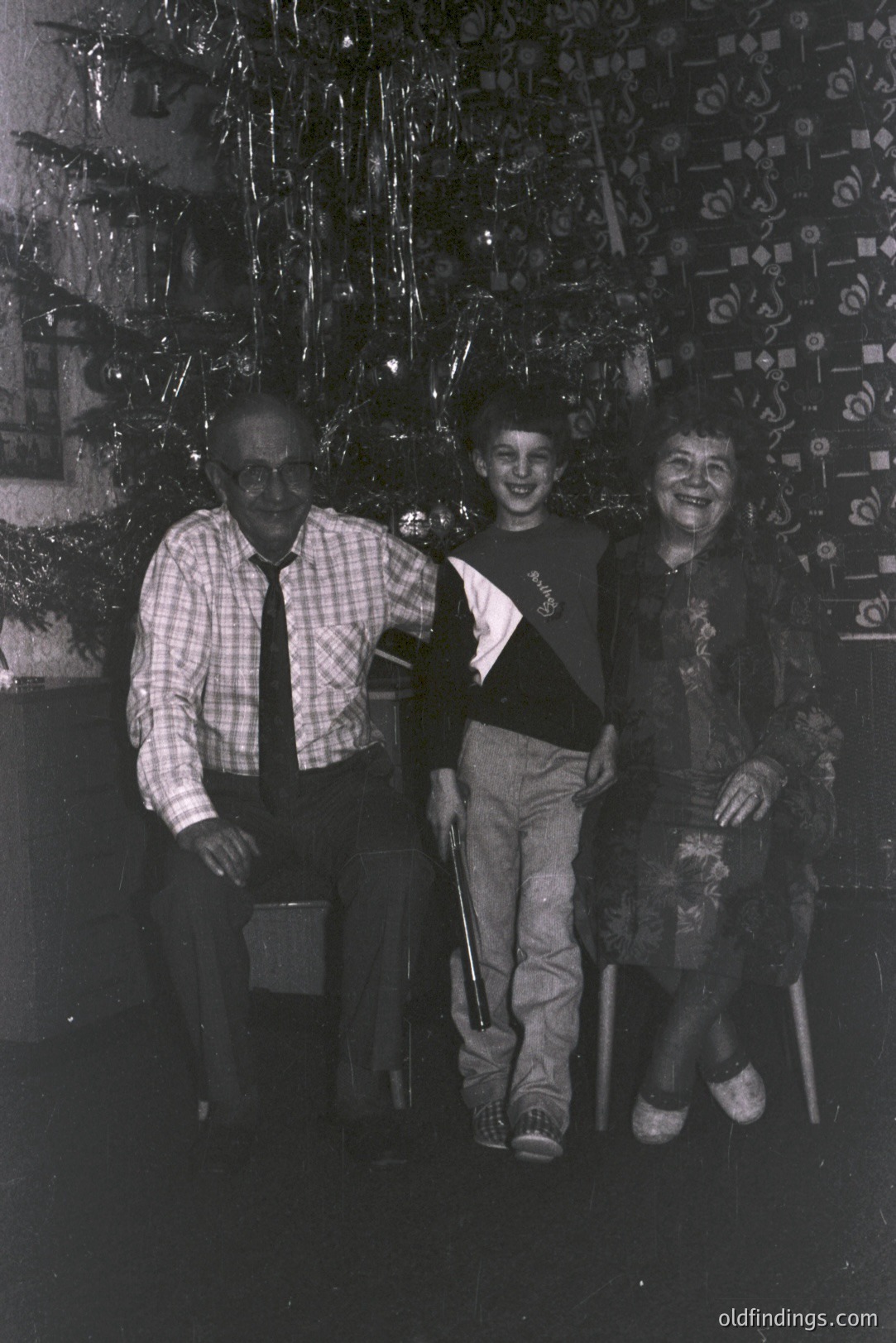 A multigenerational portrait; an elderly man in plaid, a young boy, and an older woman seated near a tinsel-decorated Christmas tree. Likely 1970s family gathering, characterized by distinctive fashion trends. A poignant glimpse into domestic life. Good for genealogy & vintage aesthetic.