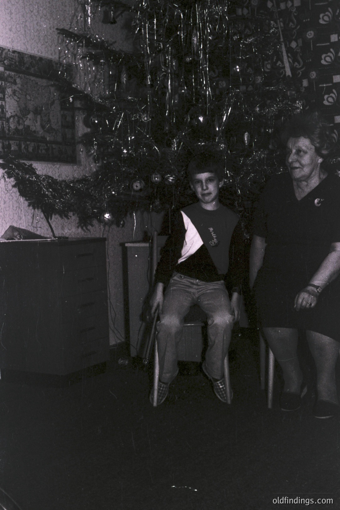Monochrome portrait of a young man seated under a heavily decorated Christmas tree. An older woman stands to his right, both appearing formally posed. Likely a family portrait from the 1970s or 1980s. Subject's clothing suggests casual style typical of the era.