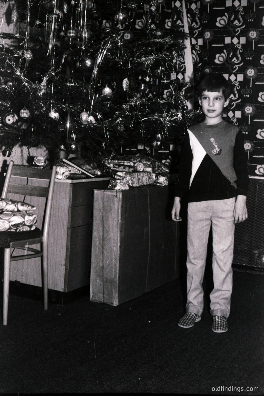 A young boy stands in front of a heavily decorated Christmas tree, surrounded by wrapped gifts. He wears a distinctive color-block sweater and plaid trousers, a style typical of the 1970s. Likely a family snapshot; valuable for nostalgic or historical design reference.