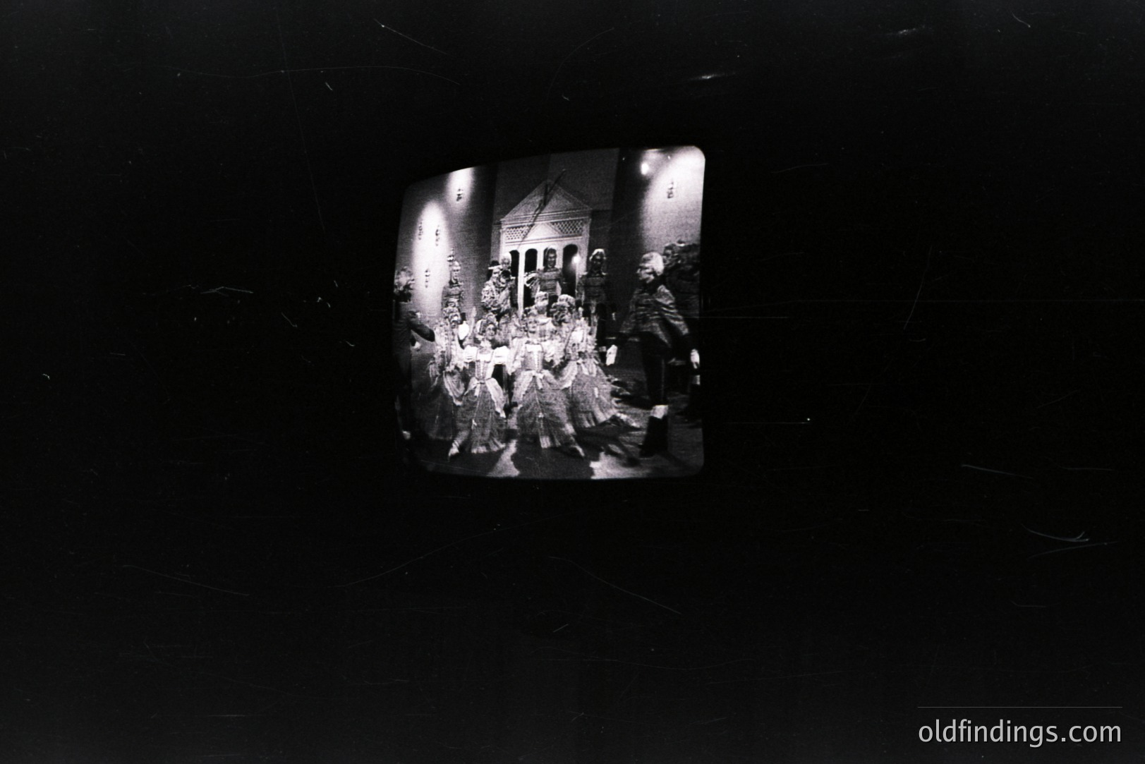 A group of elaborately costumed performers stand in a grand doorway, possibly for a stage production. Intricate masks and feathered headdresses are prominent. Architectural detail suggests a theatrical set or a historic building façade. Appears to be a still from vintage film. Likely 1970s or 80s era.