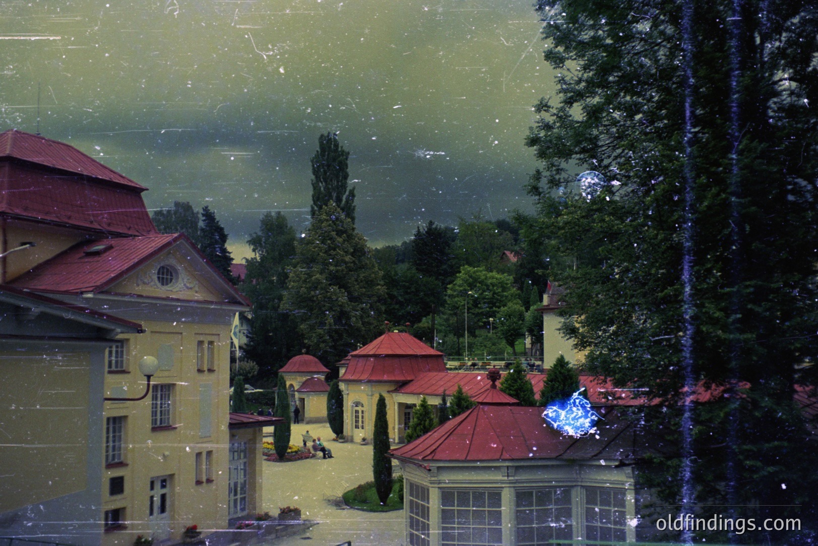 Elegant, multi-story buildings with red tile roofs define a grand courtyard, likely a historic hotel or estate. Lush greenery and a dark, overcast sky complete the scene. Architecture suggests Central/Eastern European influence, potentially 1930s-1960s. Photograph shows signs of age/damage.