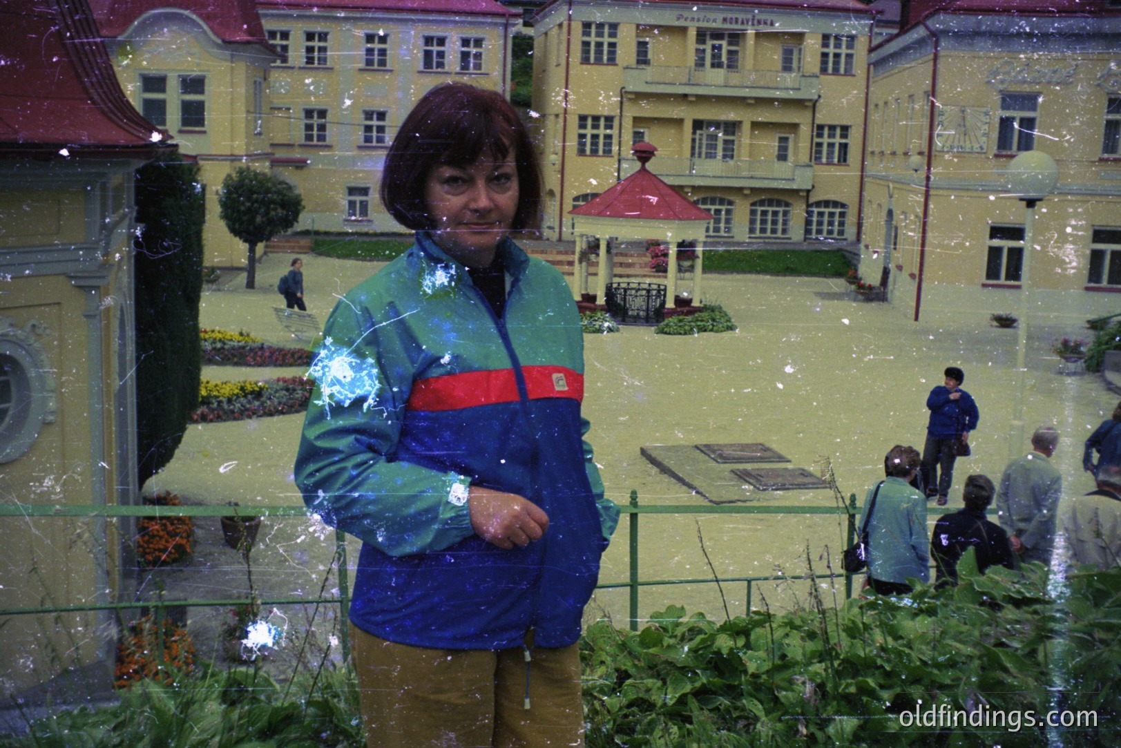 A woman in a blue and green tracksuit stands in front of a miniature alpine village, with a fountain in the center and buildings lining the perimeter. The scene is staged, appearing underwater with artificial snow. Likely a tourist attraction. 1980s-1990s.