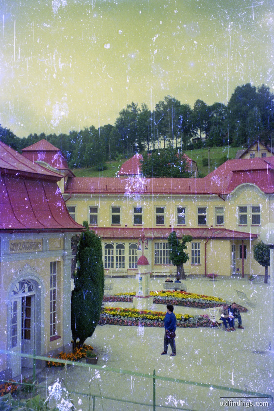 Elegant, symmetrical architecture with a terracotta tiled roof; likely a grand spa or hotel. A tiered, colorful flowerbed anchors the courtyard space. A solitary figure walks amidst greenery & distant hills. Appears to be a vintage photograph with signs of age.