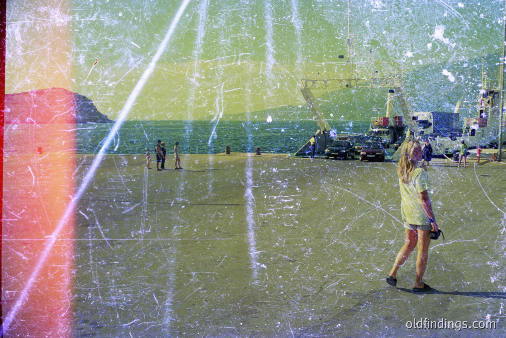 A woman in a yellow shirt walks along a concrete expanse, likely a port or industrial area, with a seaside backdrop. Shipping cranes and vehicles are visible. The image exhibits significant film grain and scratching, suggesting age or deliberate aesthetic effect. Possibly 1970s.