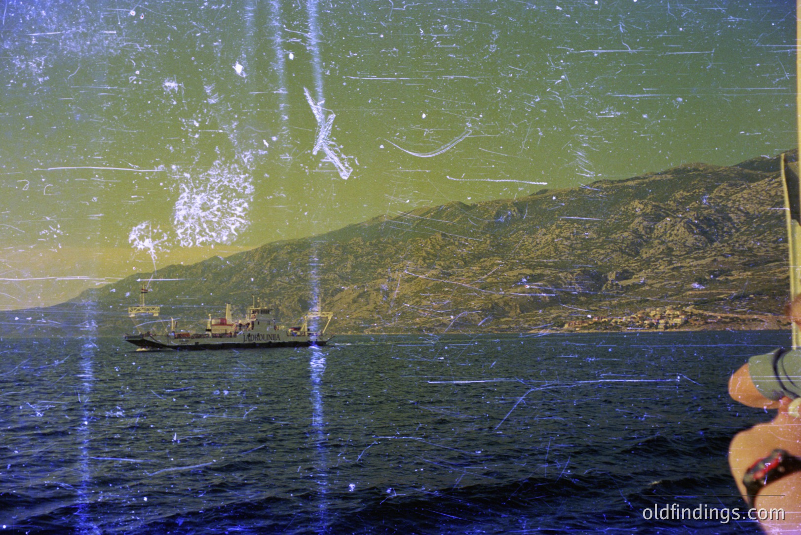 A cargo or ferry ship transits a coastal waterway. Rugged, forested mountains rise in the background with a glimpse of a town at their base. Visible signs of age and wear exist on the photograph; film grain and surface scratches are apparent. Likely captured in the 1970s or 1980s.