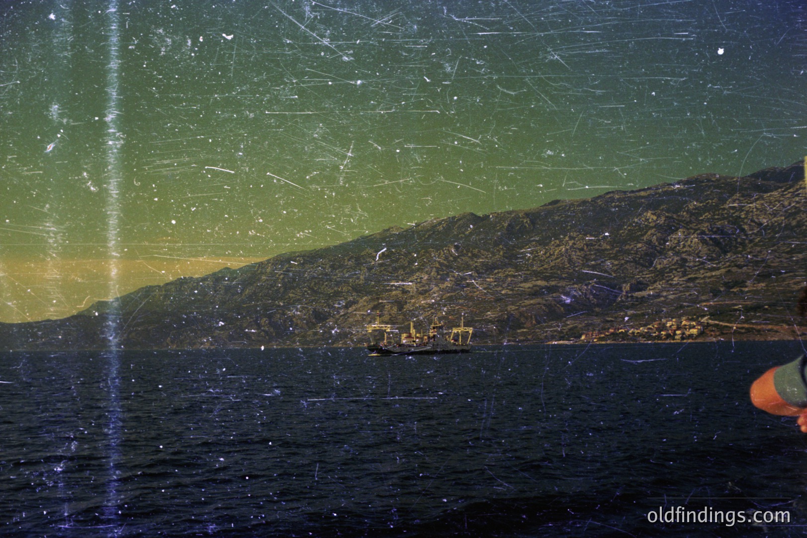 A cargo ship at anchor in a dark bay, framed by a rugged, mountainous coastline with visible lights from a coastal town. Likely a port or industrial area. Strong vertical scratches across the frame indicate age/storage damage. Film aesthetic suggests the 1970s.