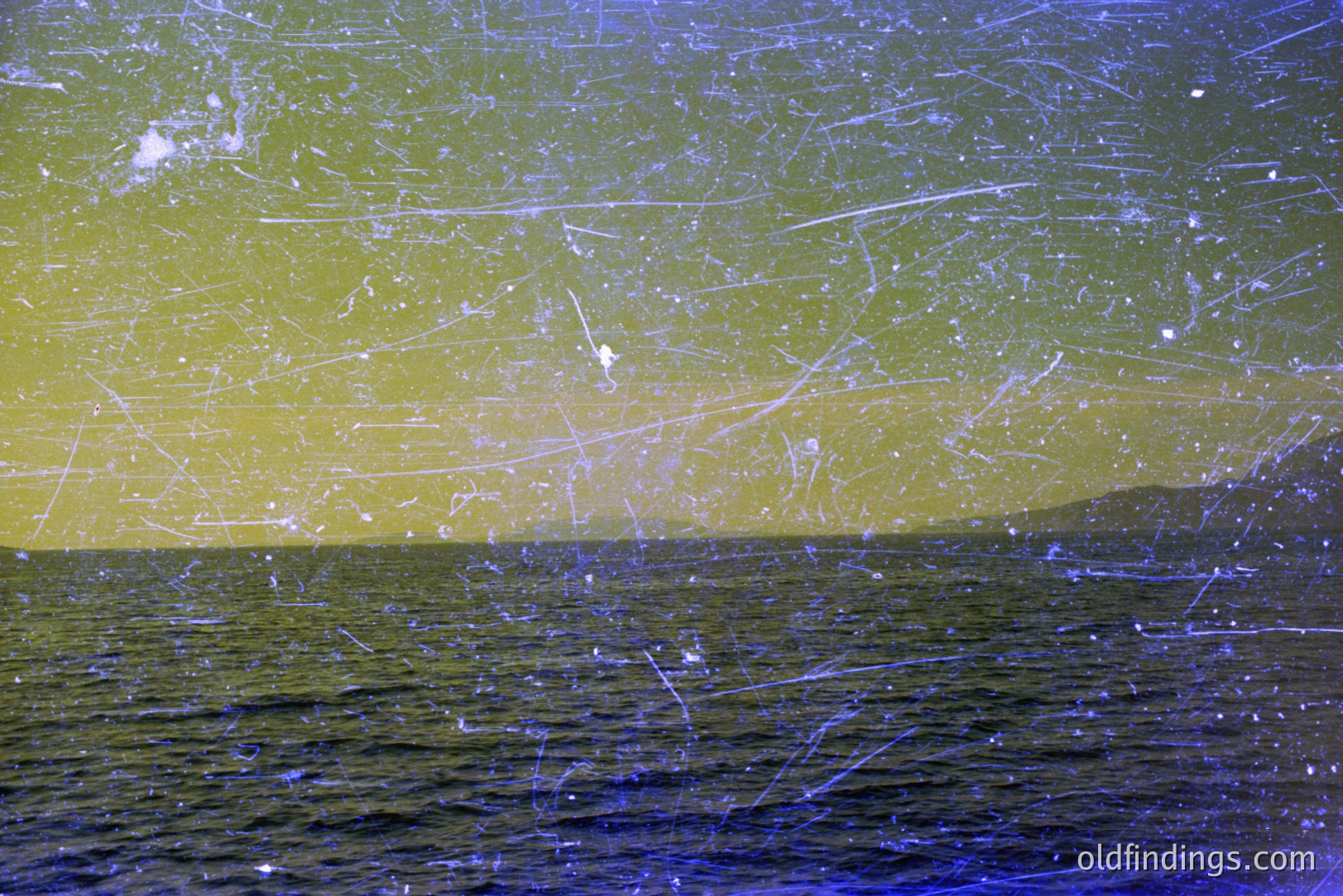 Seascape with gentle waves and distant, low-lying landmass. Heavily scratched/damaged film shows extensive surface wear, indicative of age and storage conditions. Likely a holiday snapshot, potentially from the 1960s-1970s. Color palette features blues, yellows, and hints of green.