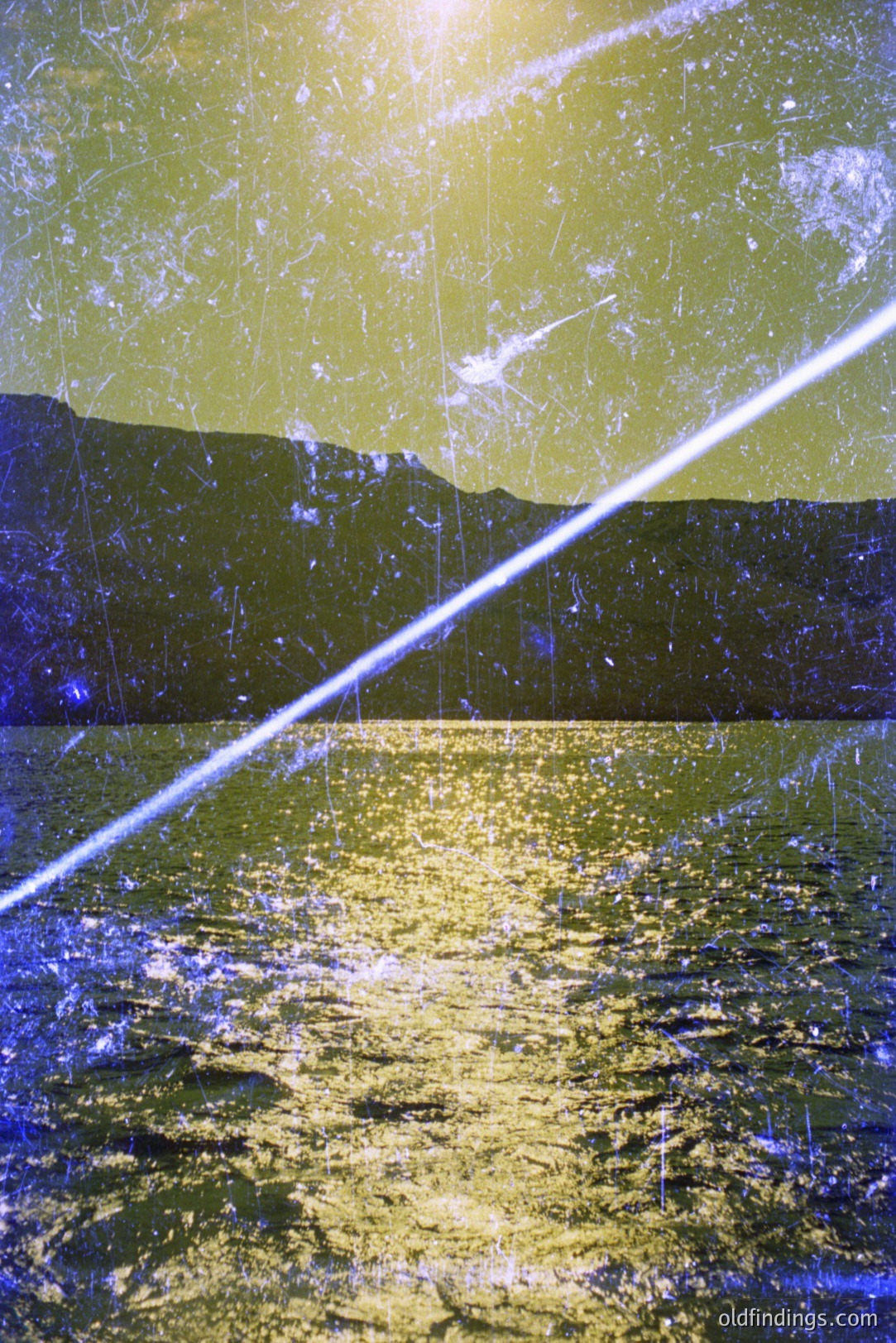 A wide, diagonal lens flare dominates this landscape photo. Shows a calm body of water reflecting sunlight, framed by a hazy mountain range in the background. Shows signs of age and photographic degradation, including dust and scratches. Likely a mid-century snapshot.