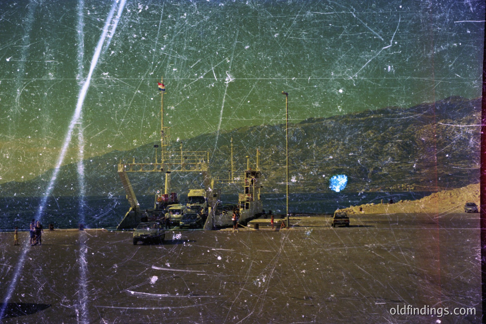 Seaside scene features a landing craft carrier and personnel unloading cargo. The coastal landscape includes a beach, vehicles, and distant hills. Likely 1970s or 80s based on film quality and equipment. Potential utility for stock, historical research, or design referencing a military logistical operation.