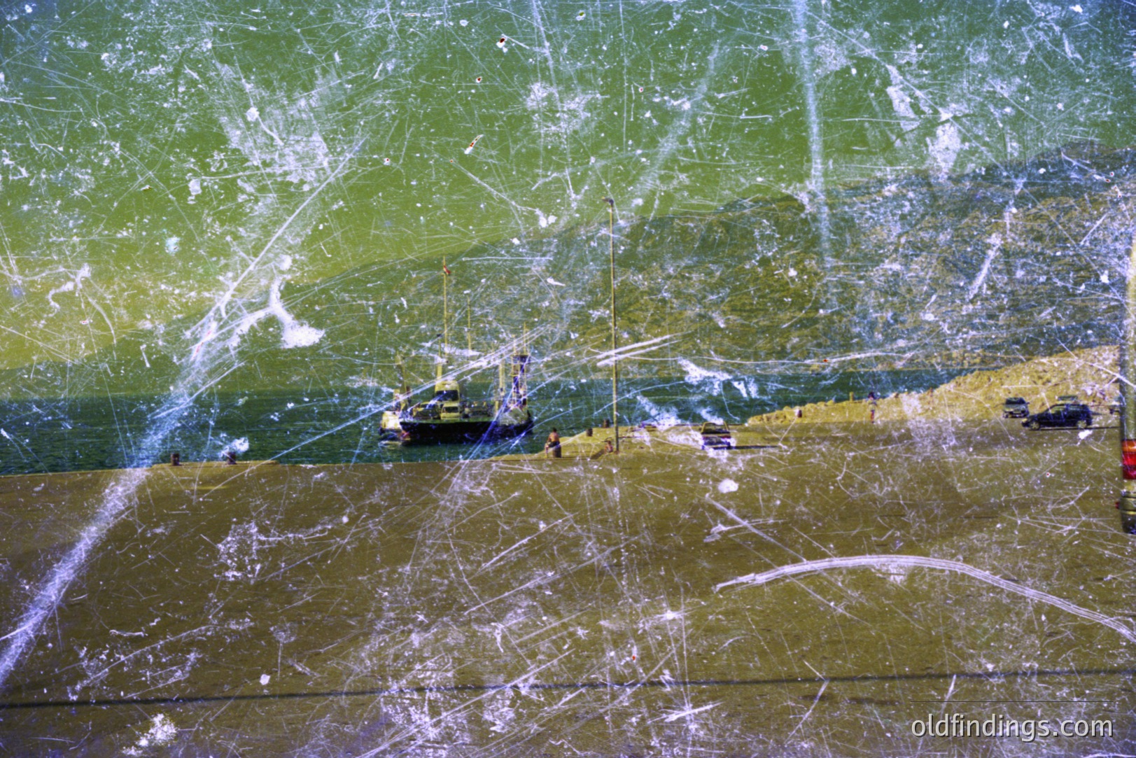 A coastal view, likely a beach or harbor, featuring a small fishing boat moored near the shore. The terrain slopes upwards, covered in vegetation. Visible wear and scratches suggest age/storage issues. Appears to be a vintage color photograph, possibly from the 1970s.