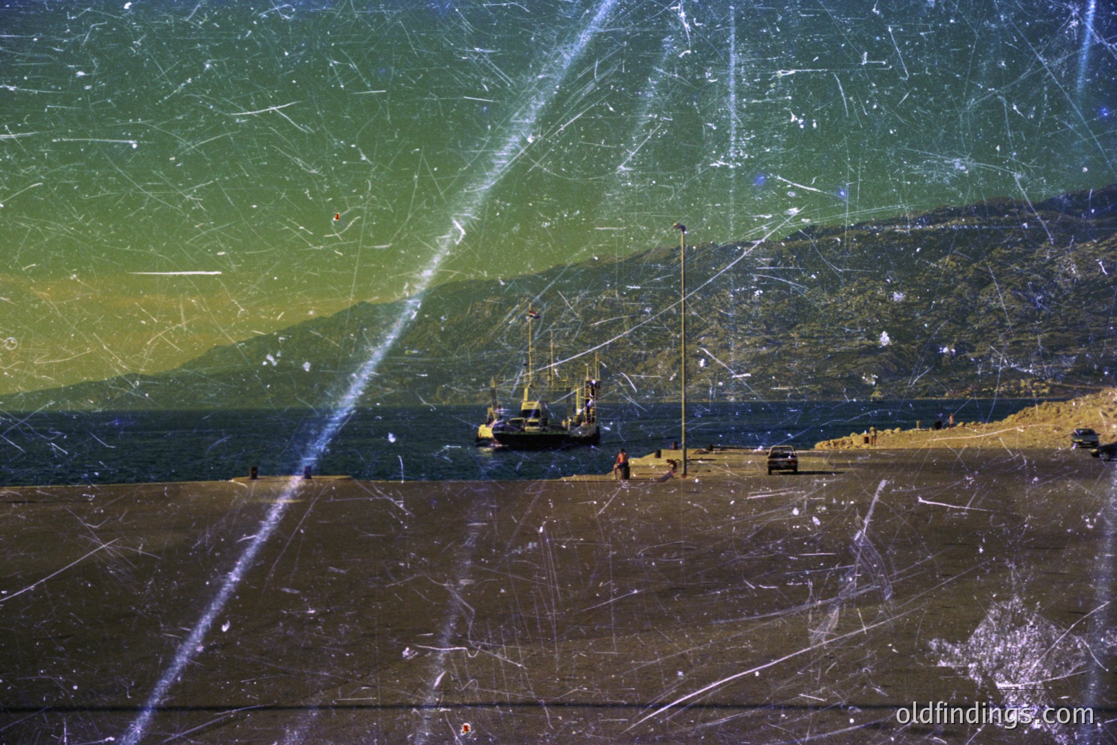 Seascape featuring a working dock, likely a port or industrial area. Visible are cranes, a small vessel, and what appears to be a construction site. Sloping hills meet the water's edge. Strong vertical scratches across the image suggest age and possible film damage.