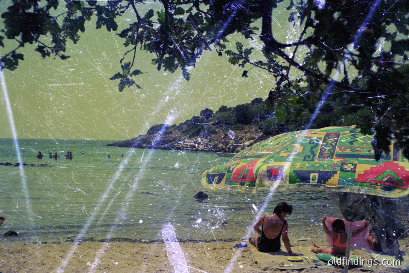 A vintage coastal scene: two sunbathers relax on a sandy beach with turquoise water & a rocky headland visible in the distance. A patterned beach umbrella provides shade. Likely taken in the 1970s, showing signs of film grain & light leaks.