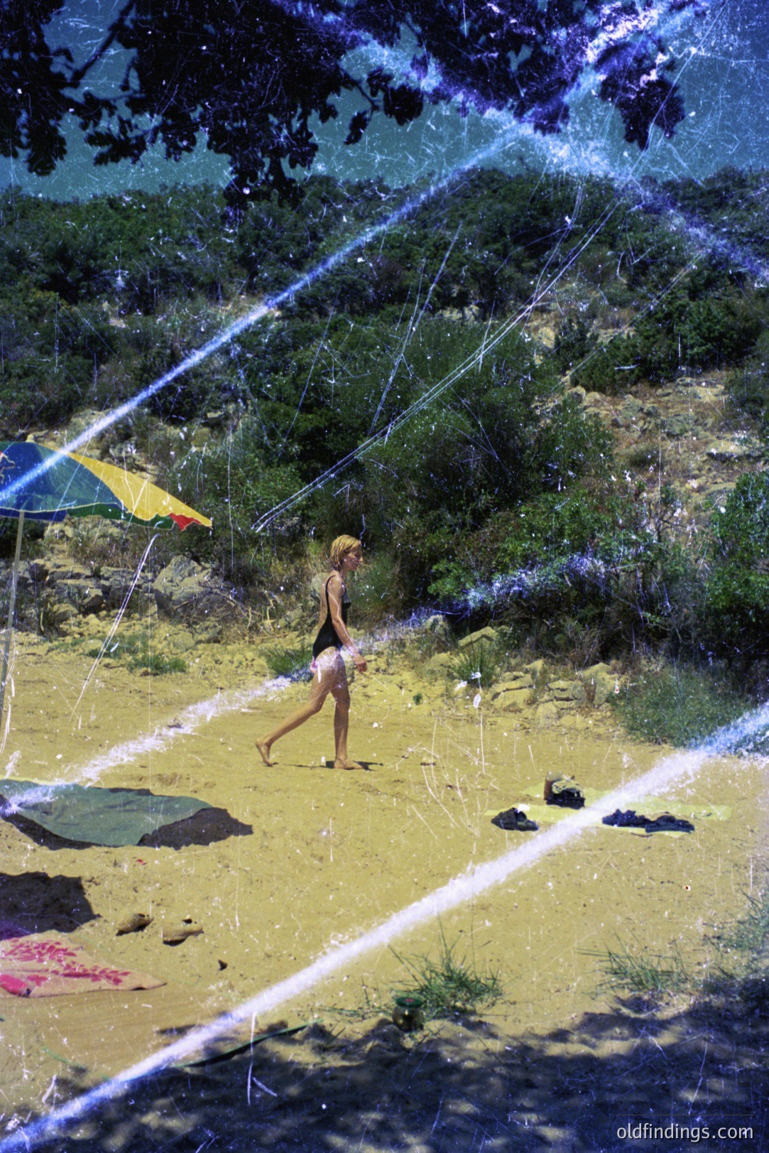A young woman walks along a sandy beach with yellow-sand dunes & verdant foliage in background. A striped beach umbrella sits nearby, & towels are strewn on the sand. Visible film grain & significant surface scratches evoke a vintage feel. Likely 1970s or 80s.