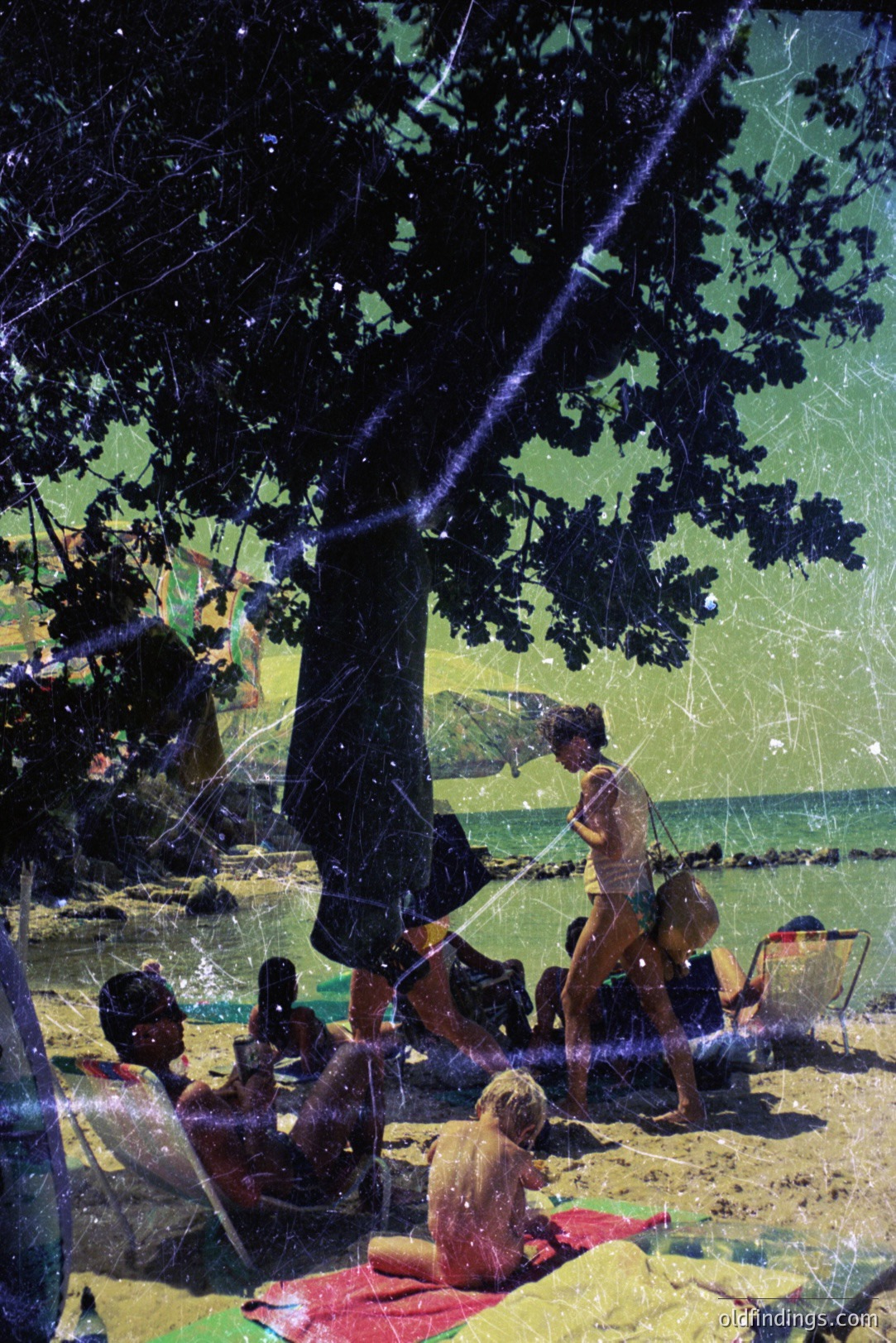 Bathers relax on a sandy beach beneath a large tree, overlooking a turquoise sea. A young woman in a bathing suit poses playfully. Visible are beach chairs, towels, and a child seated on a red blanket. The image shows visible film grain and wear. Likely a resort scene, 1960s-1970s.