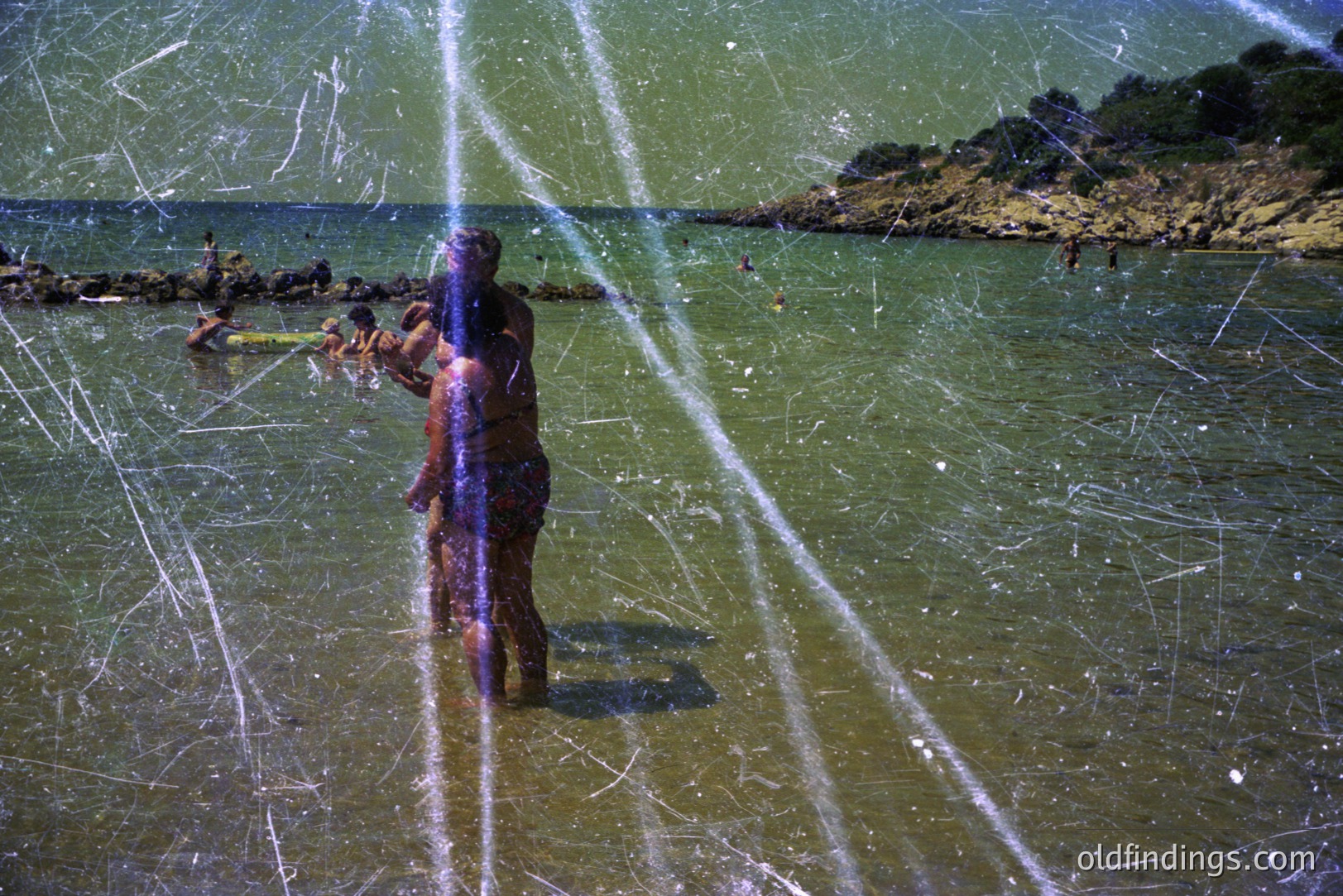 A color photo captures two figures wading in shallow, clear water; one appears to be holding the other. Background shows a rocky shoreline and sparse vegetation. Apparent light leaks dominate the frame, indicating age/film damage. Likely taken in the 1970s, coastal location, perhaps Southern Europe.
