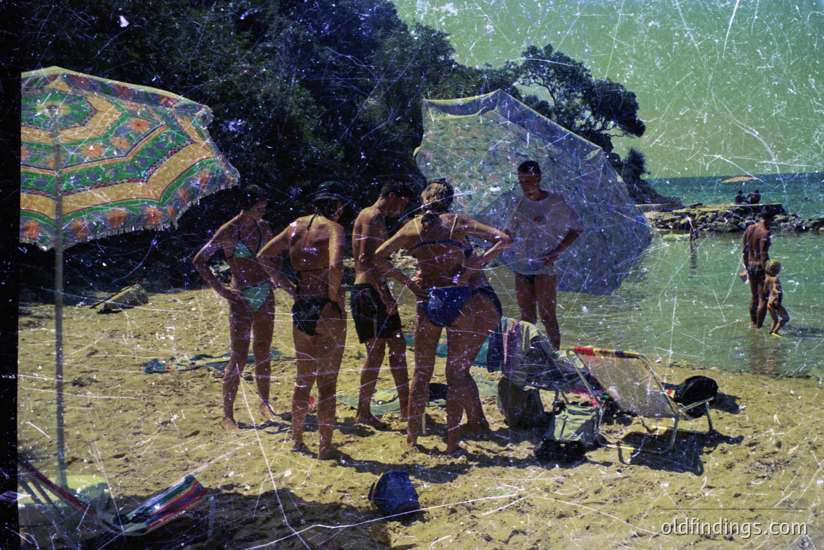 A group of six young people on a sandy beach, seemingly mid-conversation. Visible swimsuits & vacation attire suggest a leisure setting, likely 1960s-1970s. An umbrella and beach gear add to the scene. Photograph exhibits age-related wear (scratches).