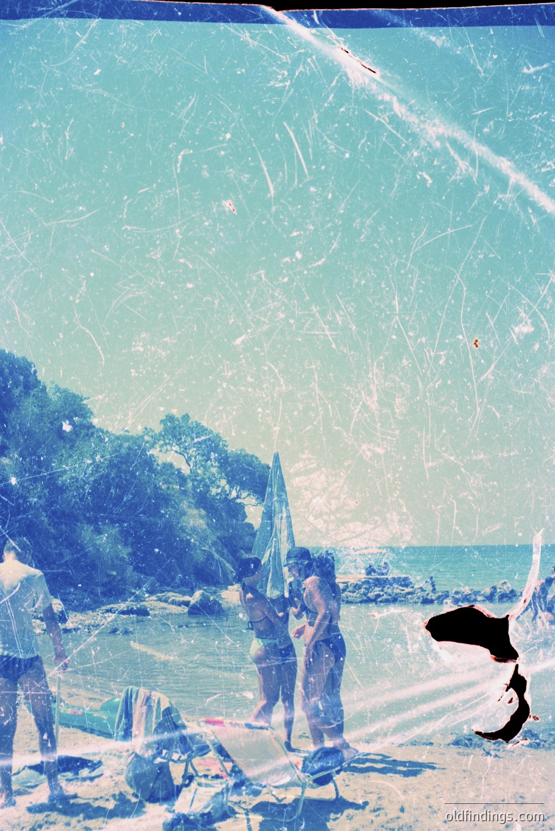 A candid seaside scene, likely from the 1960s or 70s. Three figures in swimwear stand near the shoreline, sheltered by a striped umbrella. Distant rocky headlands define the coast. The photo exhibits significant film grain & scratches, contributing to a vintage aesthetic. Potentially Bulgaria, Varna.