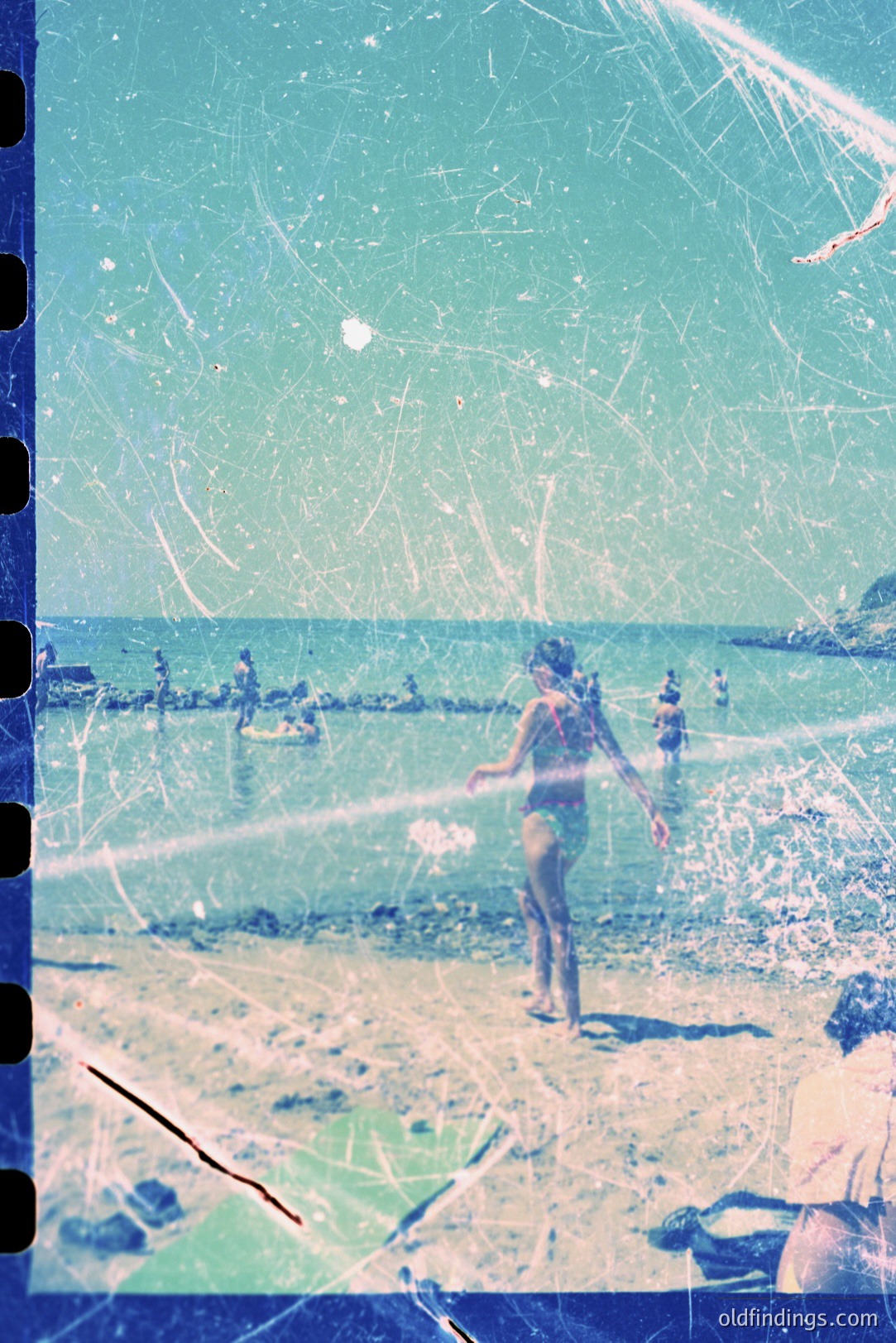 A youthful woman in a swimsuit wades into turquoise water on a sandy beach. Other figures are visible in the distance along the shoreline. Image shows signs of age – scratches and dust visible on the film. Likely a candid, summertime snapshot, c. 1960s-1970s.