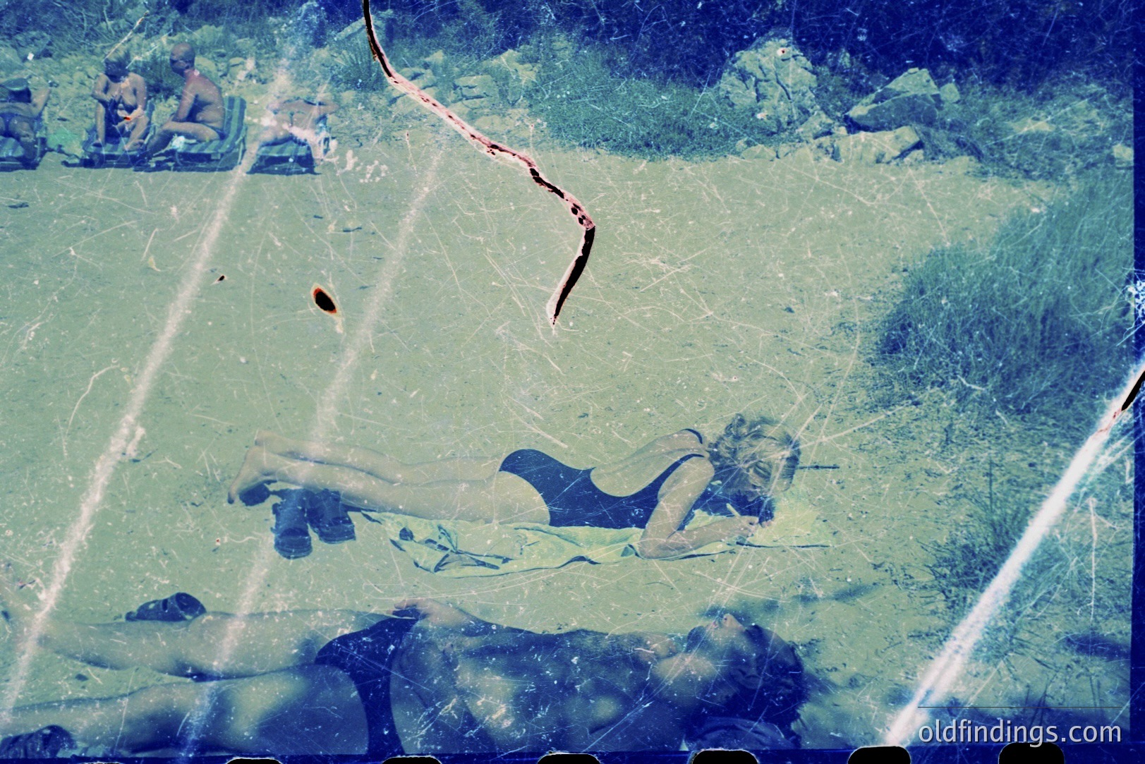 A vintage color film captures two women sunbathing on what appears to be a beach. One woman reclines, while the other is positioned above her. Sparse vegetation and blurred background suggest a coastal environment. The image displays visible film grain and scratches, indicating age. Likely 1960s or 1970s.