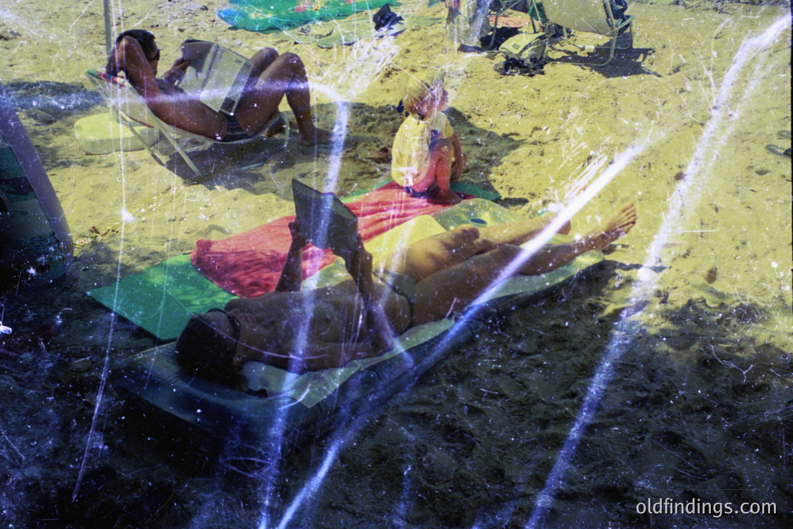Sunbathers relax on a sandy beach, partially obscured by a double exposure effect showing rain or water spray. Likely a coastal resort setting. The image exhibits signs of age, including visible film grain and scratches. Captures leisure and vacation atmosphere. Likely 1970s or 1980s.
