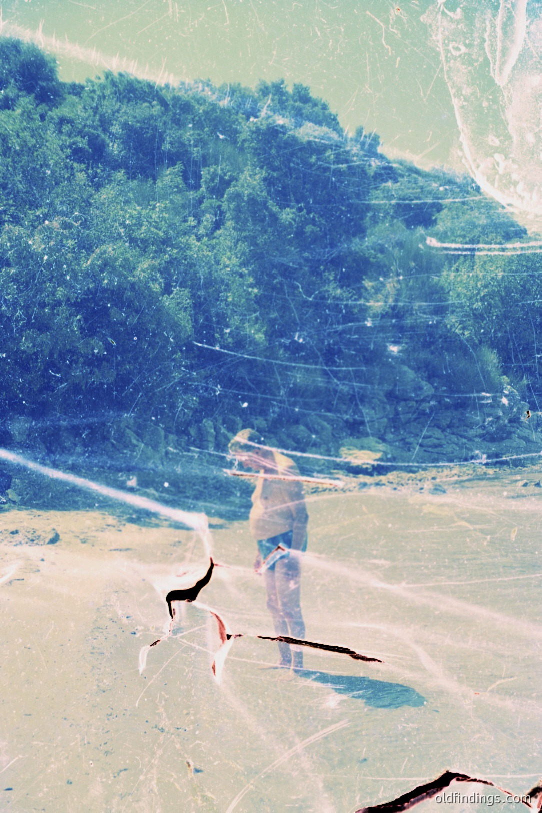 A figure stands on a beach, facing a lush, forested hillside, with water visible in the background. The image exhibits significant damage - scratches and discoloration suggesting age and possible film deterioration. Likely captured in the 1970s, possibly seaside vacation photography. Photographic value: historical ephemera, textural reference.