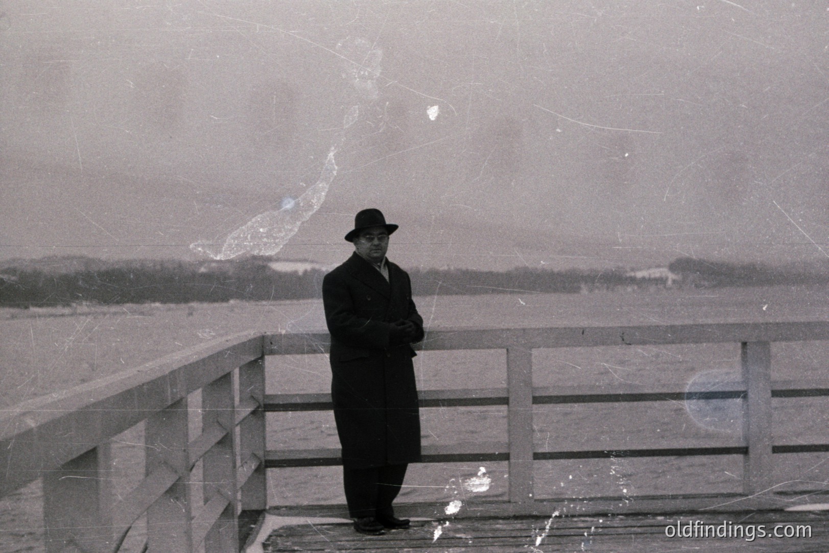 A man in a long coat and hat stands on a raised boardwalk overlooking a body of water, possibly the Black Sea. The weathered wooden structure and gentleman's attire suggest a 1960s-1970s aesthetic. Appears to be in Bulgaria, possibly near Varna. Excellent example of mid-century portraiture.