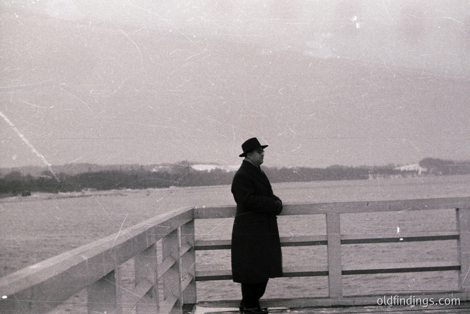 Man in dark coat and hat stands at a stone railing overlooking a body of water. Minimalist composition, likely seaside location. Possible Baltic Sea region, 1960s-1970s. Reflective, moody aesthetic. Documenting a contemplative moment or solitary figure. May be of interest to researchers studying fashion or landscape photography.