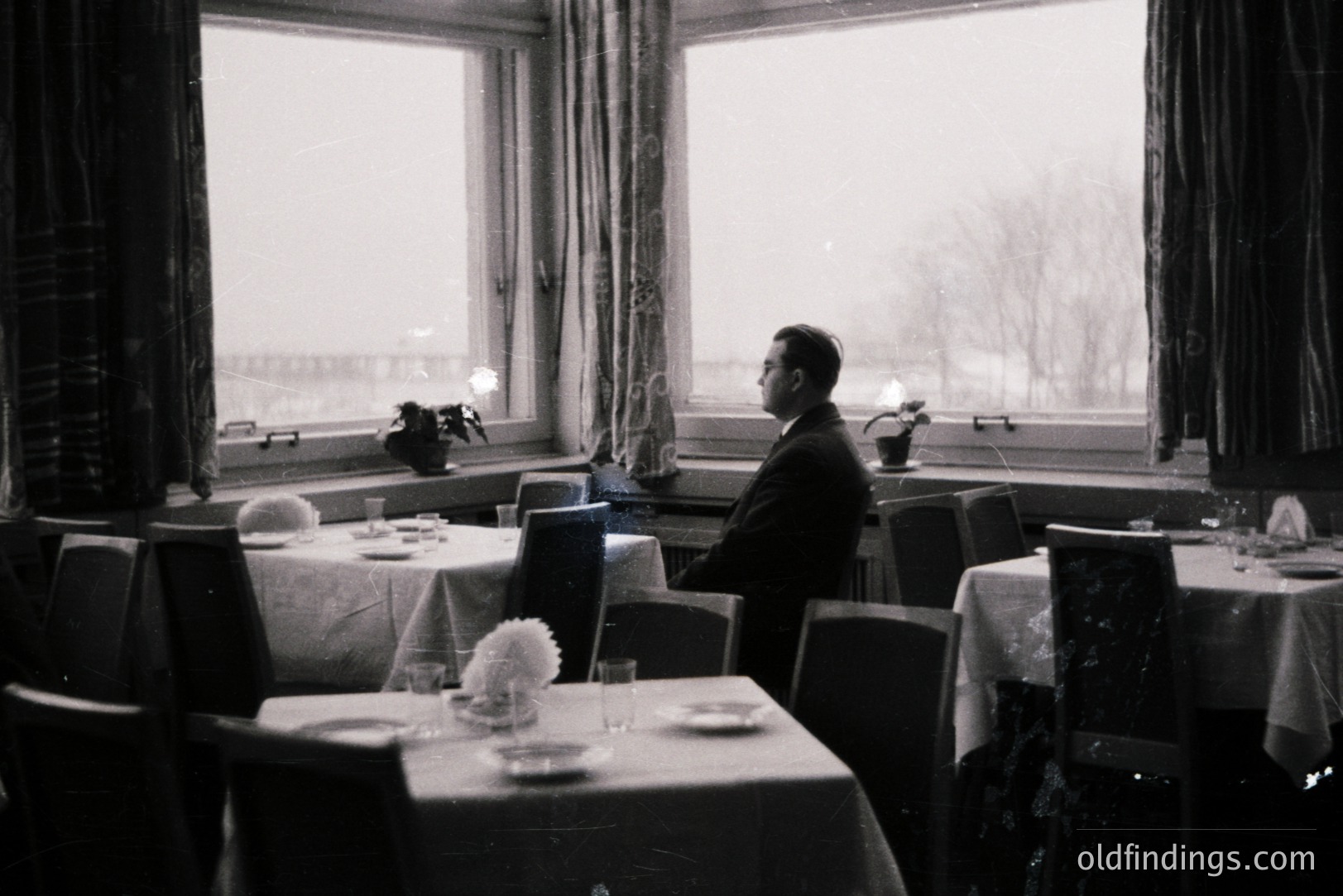 Man in suit sits in a formally set dining room, overlooking a snowy landscape. The room features draped windows, linen-covered tables, and a generally somber mood. Likely mid-20th century, possibly 1950s-1960s. Potential design reference for vintage interiors.
