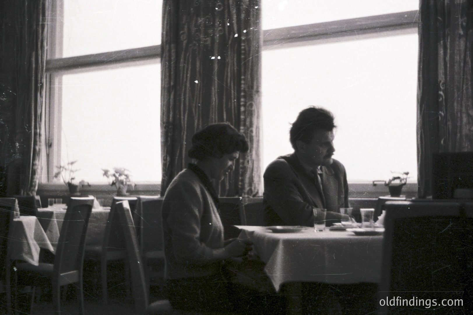 Two figures seated at a table covered with a floral tablecloth. Architectural details suggest a dining hall or restaurant setting. Likely 1960s or 1970s. Visible through the window—an unclear outdoor view. Subject matter suitable for vintage advertising or historical interiors.