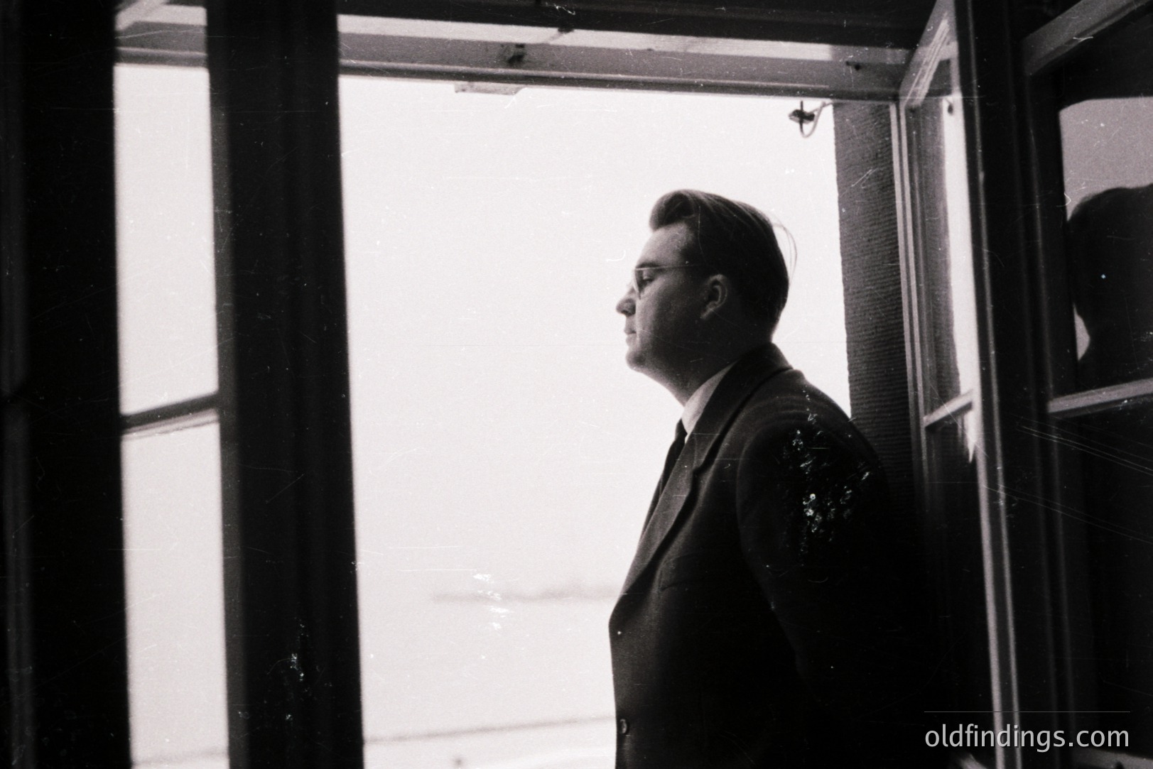 Man in a suit and glasses stands in profile near a window, gazing outward. The image, likely taken in the mid-20th century, exhibits stark contrast and grainy texture indicative of film photography. Strong architectural lines frame the subject, creating a sense of isolation. Potential stock use for mood/portraiture.