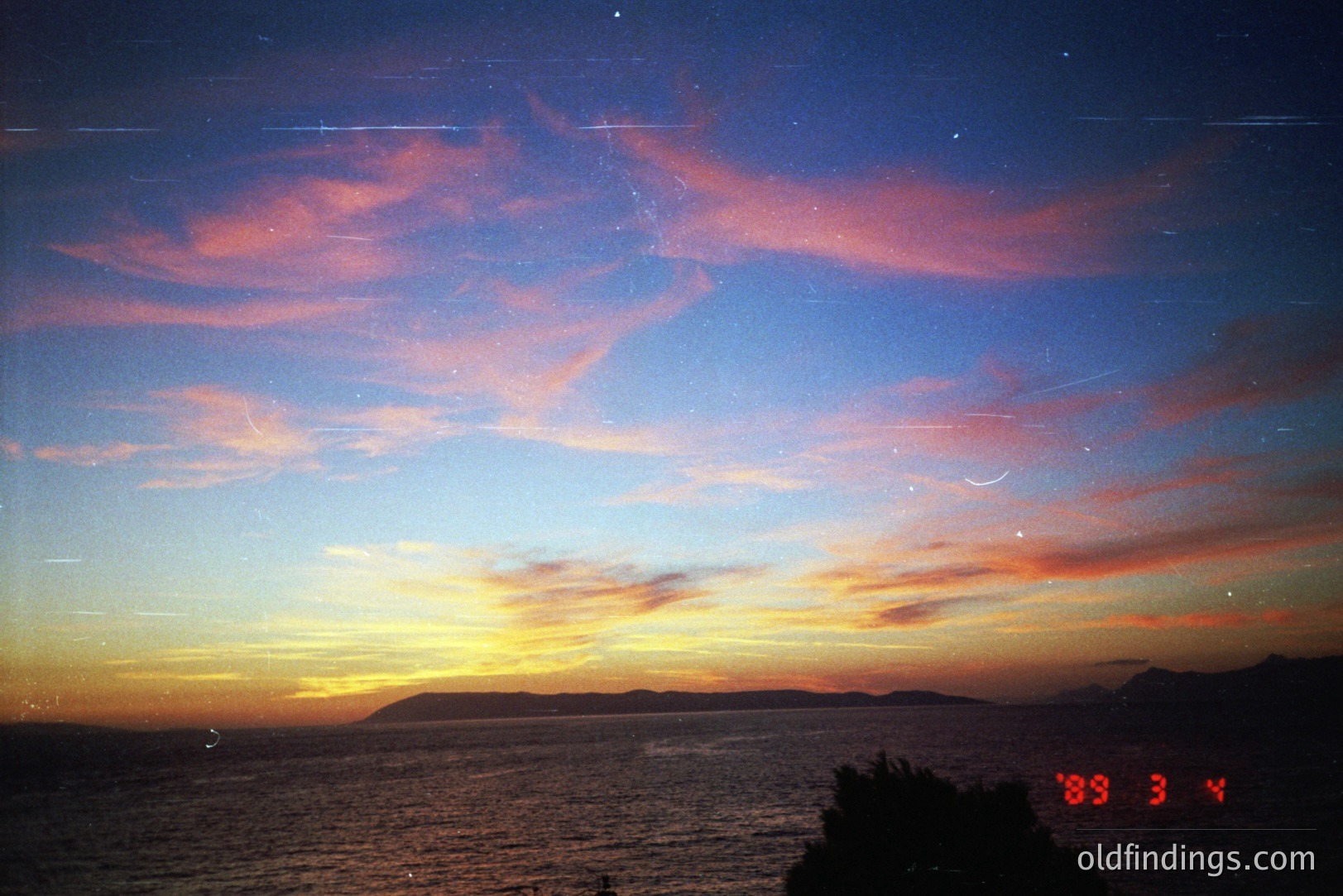 Dramatic twilight seascape with vibrant pink and orange clouds reflecting on still water. Distant landmasses silhouette the horizon. Likely coastal scene, potentially Mediterranean. Vintage film aesthetic with visible dust/light leaks.