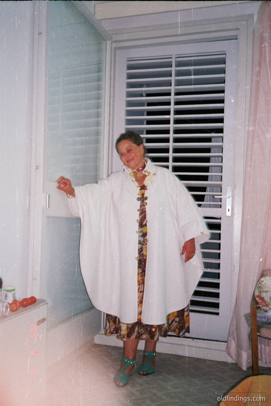 Elegant woman in a flowing white shawl stands near a shuttered window. She wears a vibrant patterned dress and green sandals, adorned with jewelry. Interior suggests a vacation home, likely 1970s-80s. A rattan chair and a ceramic lamp are visible. Likely a candid family snapshot.