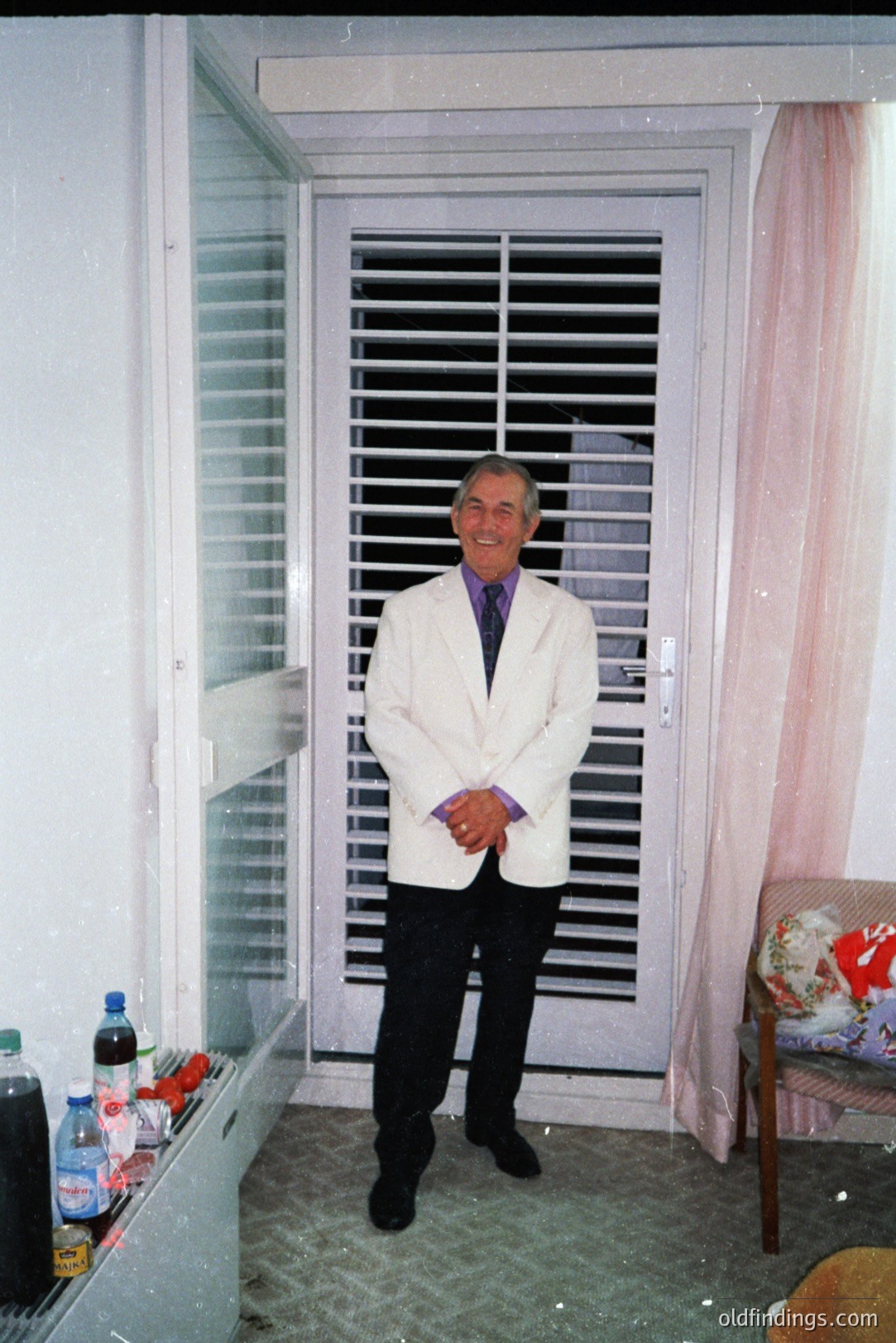 A man stands in a brightly lit room, positioned before a closed, shuttered window. He wears a white blazer, dark trousers, and a patterned purple tie. A small side table is visible, bearing bottles and assorted items. Pink curtains frame the window. Appears to be a 1970s-80s snapshot.