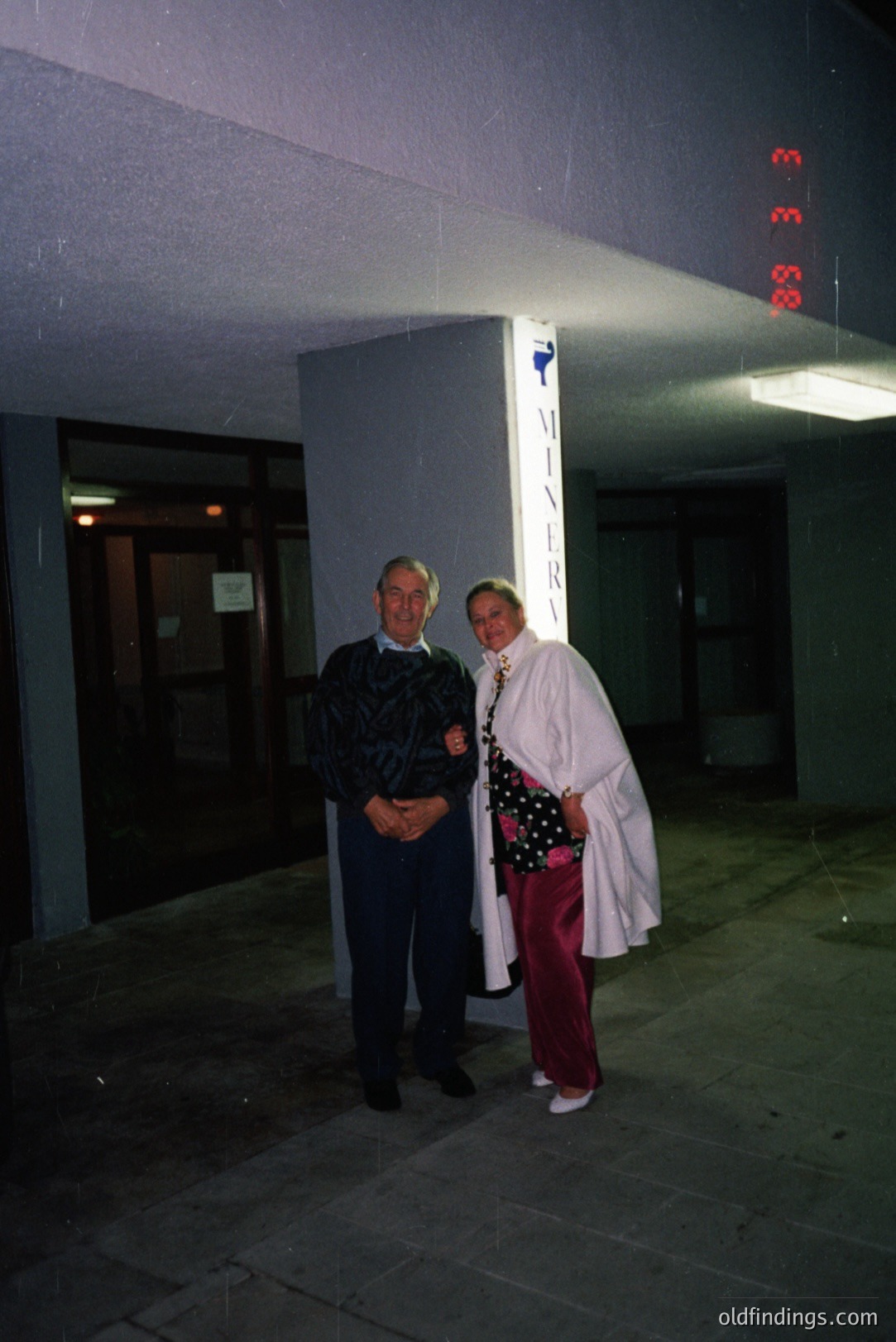 A man and woman pose outdoors near a building entrance, identifiable by a “MINERVA” sign. The man wears dark slacks and a patterned sweater; the woman, a long white coat, red pants, and a pearl necklace. Appears to be a 1980s snapshot. Architectural style suggests institutional or commercial building.