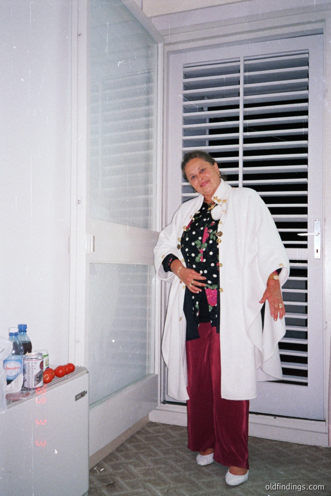 A woman stands near a window with slatted blinds, wearing a white robe, black & white polka dot top, and burgundy slacks. A mini-fridge and beverages are visible. Appears to be a hotel room or apartment interior. Likely 1980s style decor. Possible travel photograph.