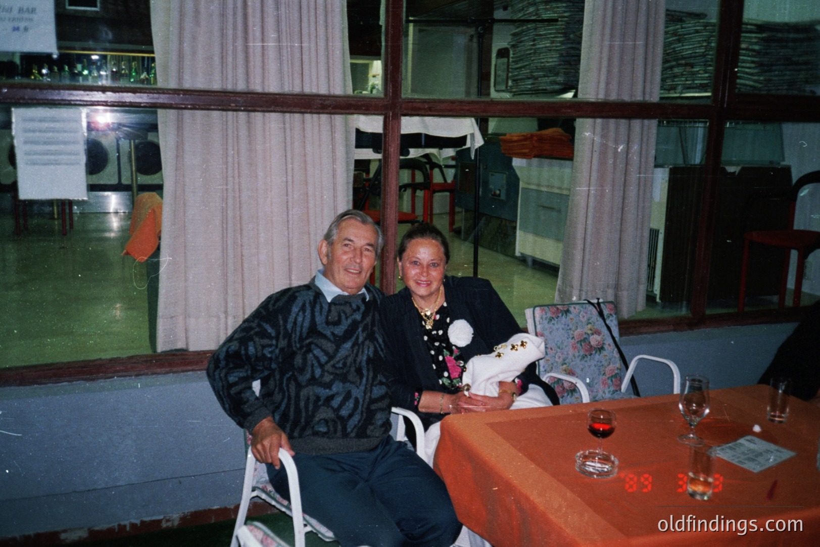 A couple poses for a candid portrait. The man, in a patterned sweater, sits in a white folding chair; the woman, in a floral-print dress, holds a plate with what appears to be bread or pastries. A red-covered table, set with glasses, is in the foreground. Interior, likely a restaurant. Appears to be late 1980s/early 1990s, based on the photo quality and attire.