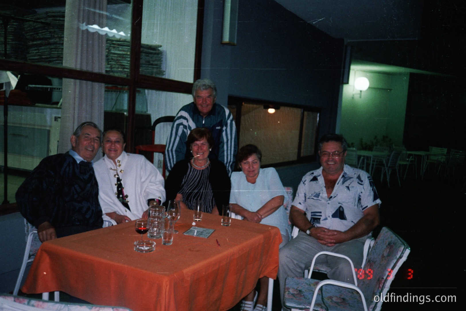 Group portrait of six adults seated around an orange-covered table, likely in a restaurant or resort setting. Visible glassware and a card deck suggest a social gathering. The décor and attire suggest a 1980s or early 1990s time period. Location and origin unknown.